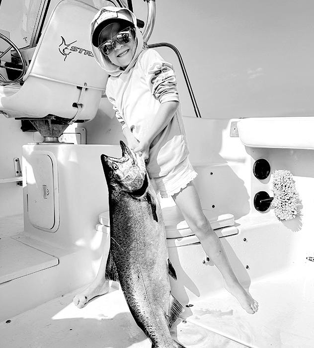 Smiling young child wearing sunglasses and a hat on a boat holding a large fish.