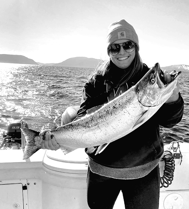 Smiling person wearing sunglasses and a beanie holding a large fish on a boat with mountains and water in the background.