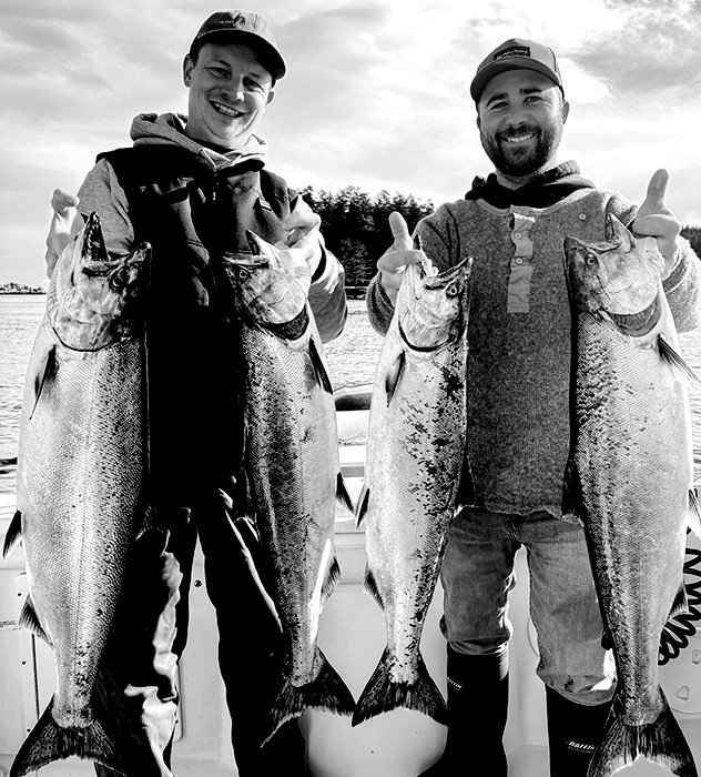 Two men standing on a boat holding large fish, smiling with water and trees in the background.