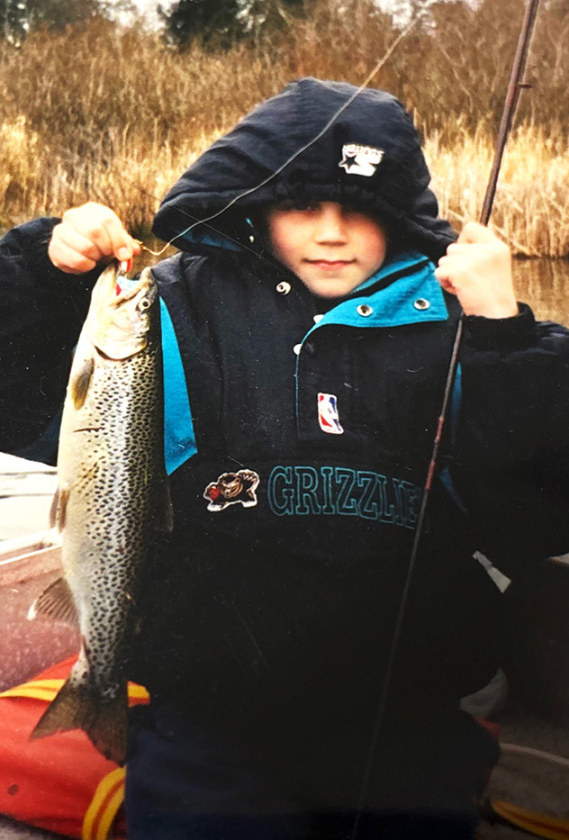 Child in a hooded jacket holding a fishing rod and a large speckled fish outdoors near water and tall grass.