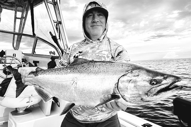 Smiling man in a hooded shirt holding a large salmon on a boat with ocean in the background.