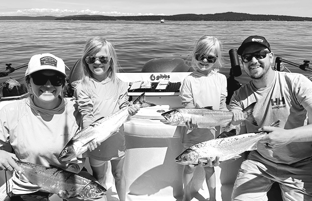 Two adults and two children on a boat holding freshly caught fish with water and distant land in the background.