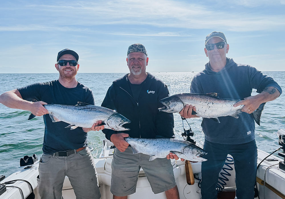 Three men on a boat holding large caught fish with the ocean and blue sky in the background.