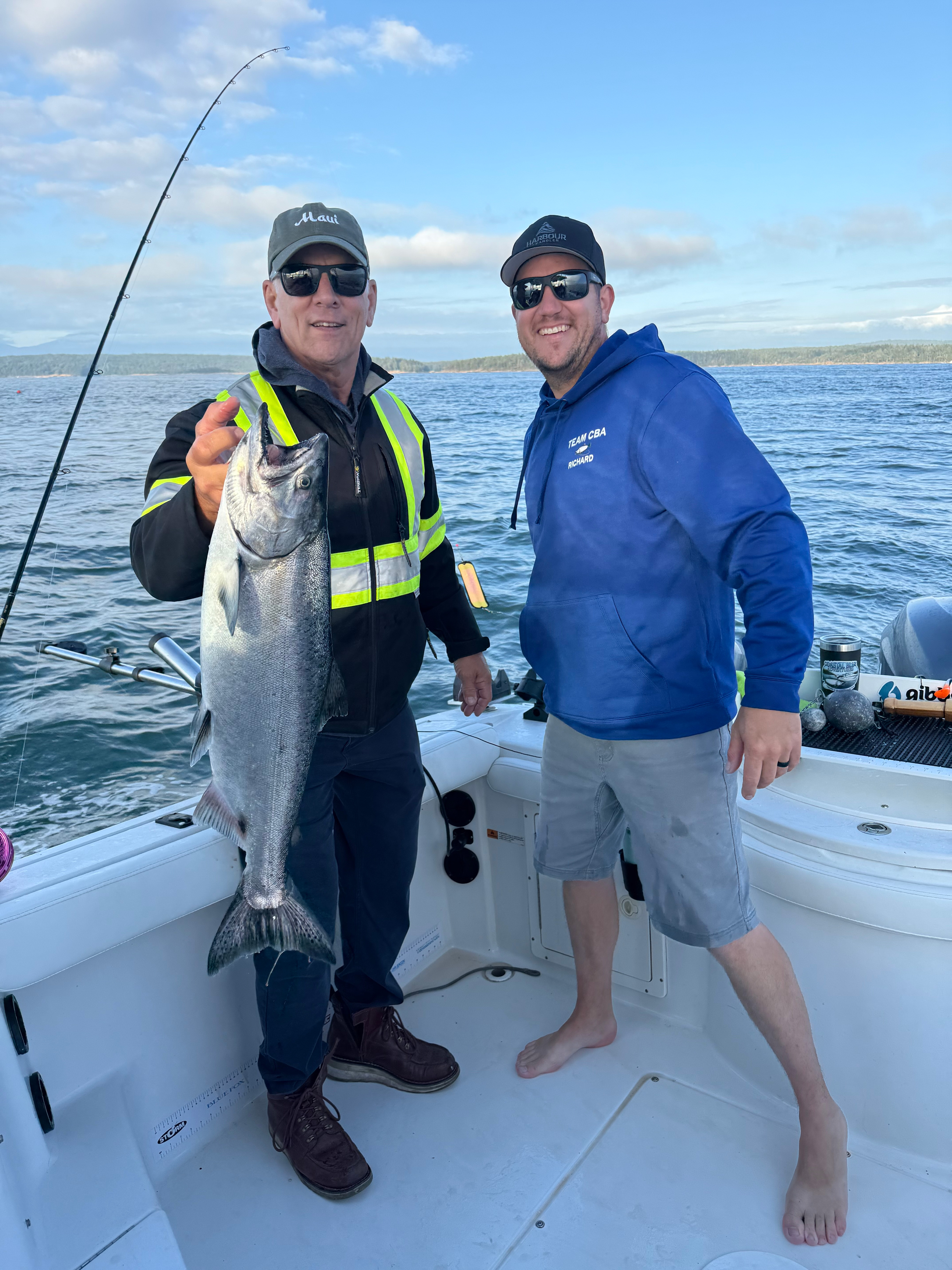Two men on a boat holding a large silver fish, with ocean and partly cloudy sky in the background.