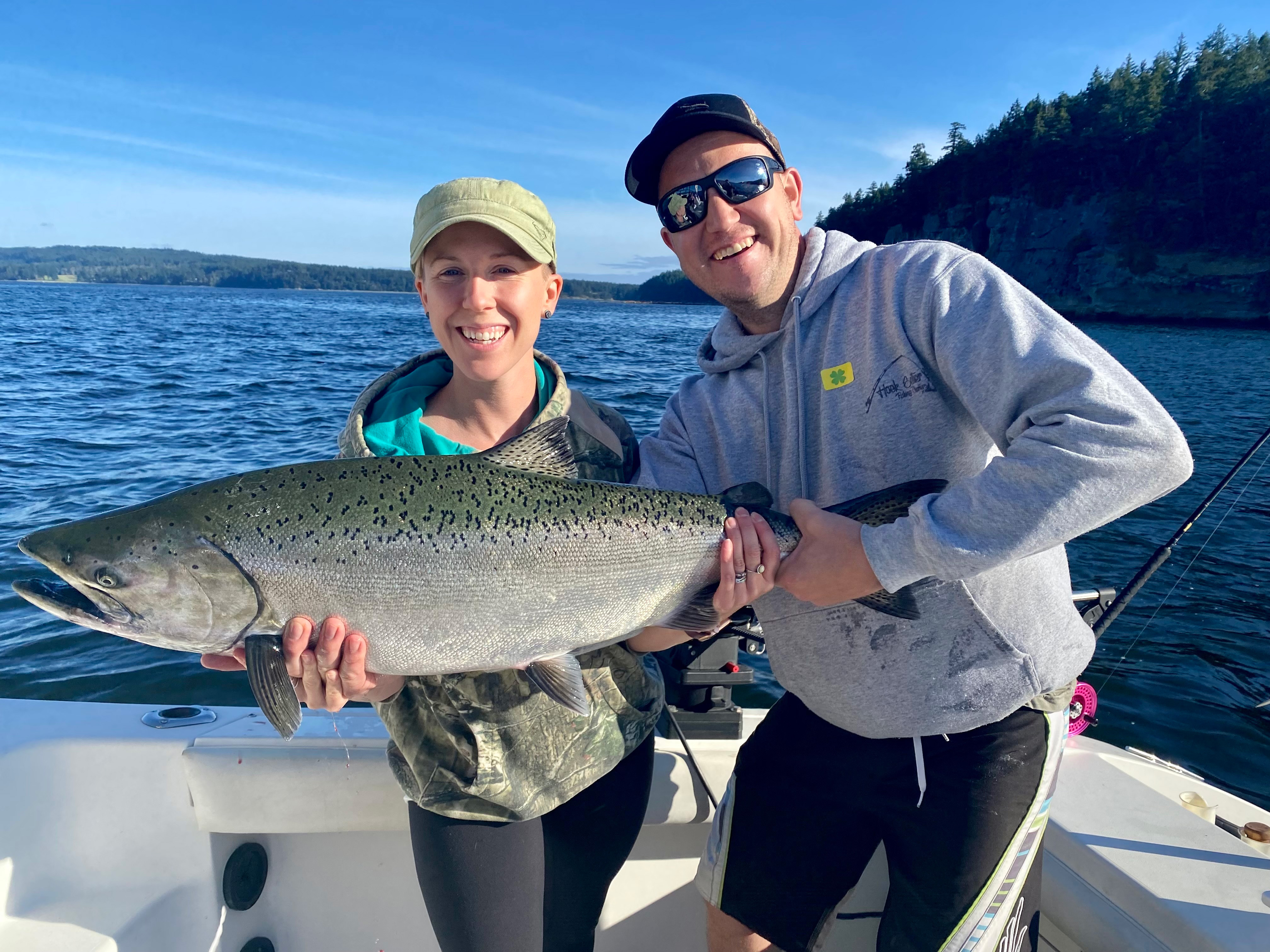 A smiling woman and man holding a large fish together on a boat with water and forested shore in the background.
