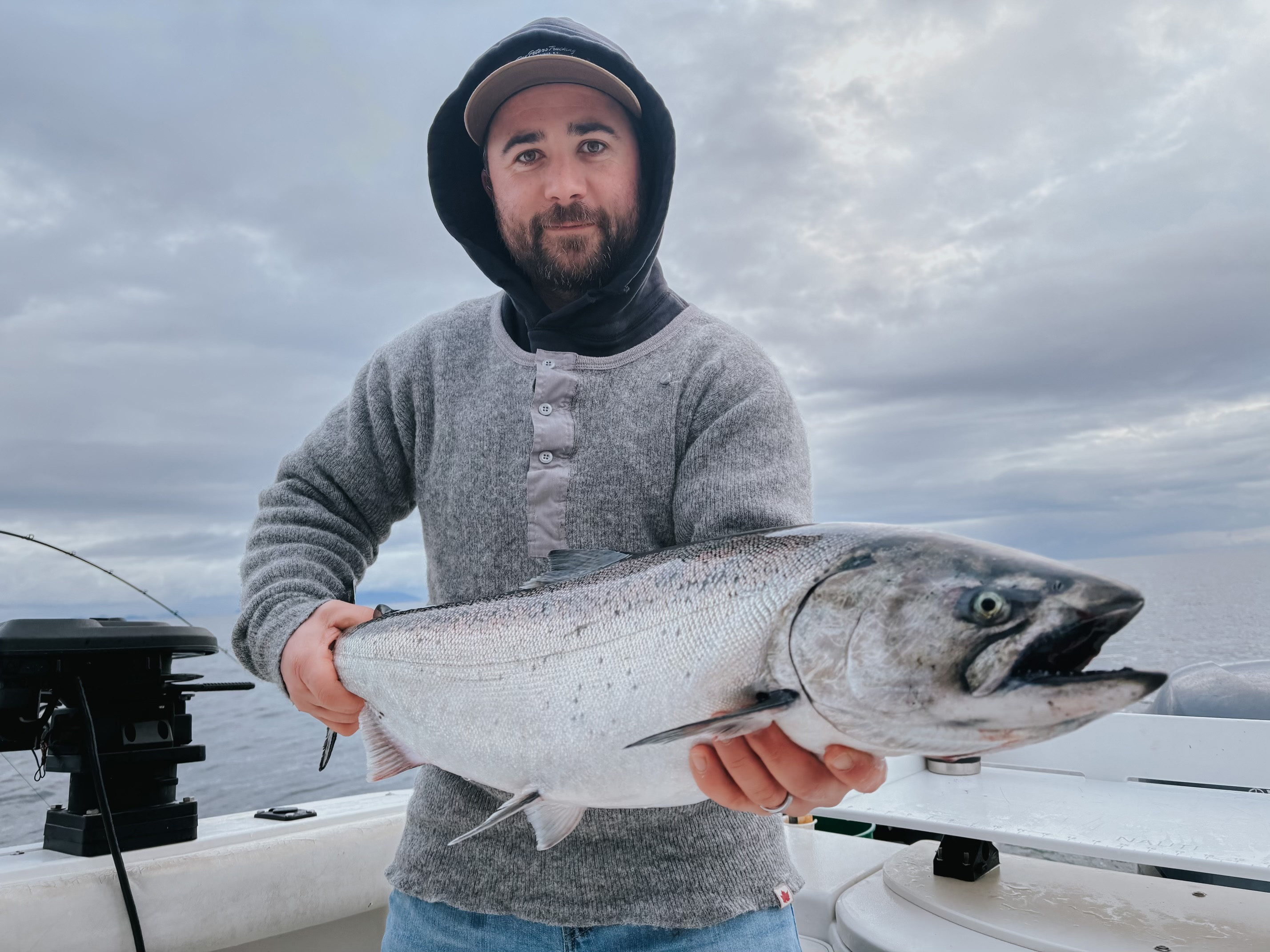 Man in a gray sweater and cap holding a large silver fish on a boat under cloudy sky.