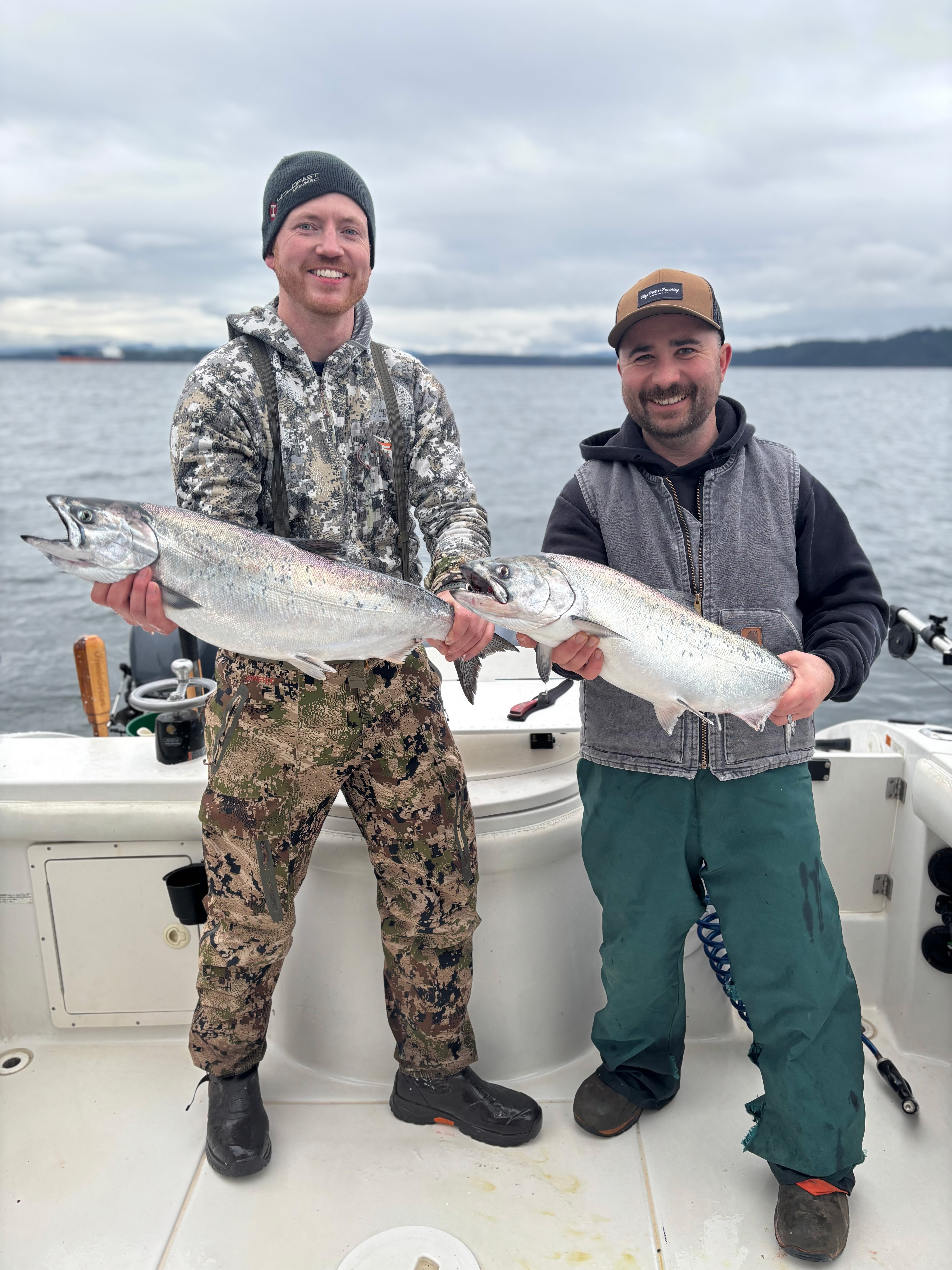 Two men smiling and holding large silver fish on a boat with water and cloudy sky in the background.