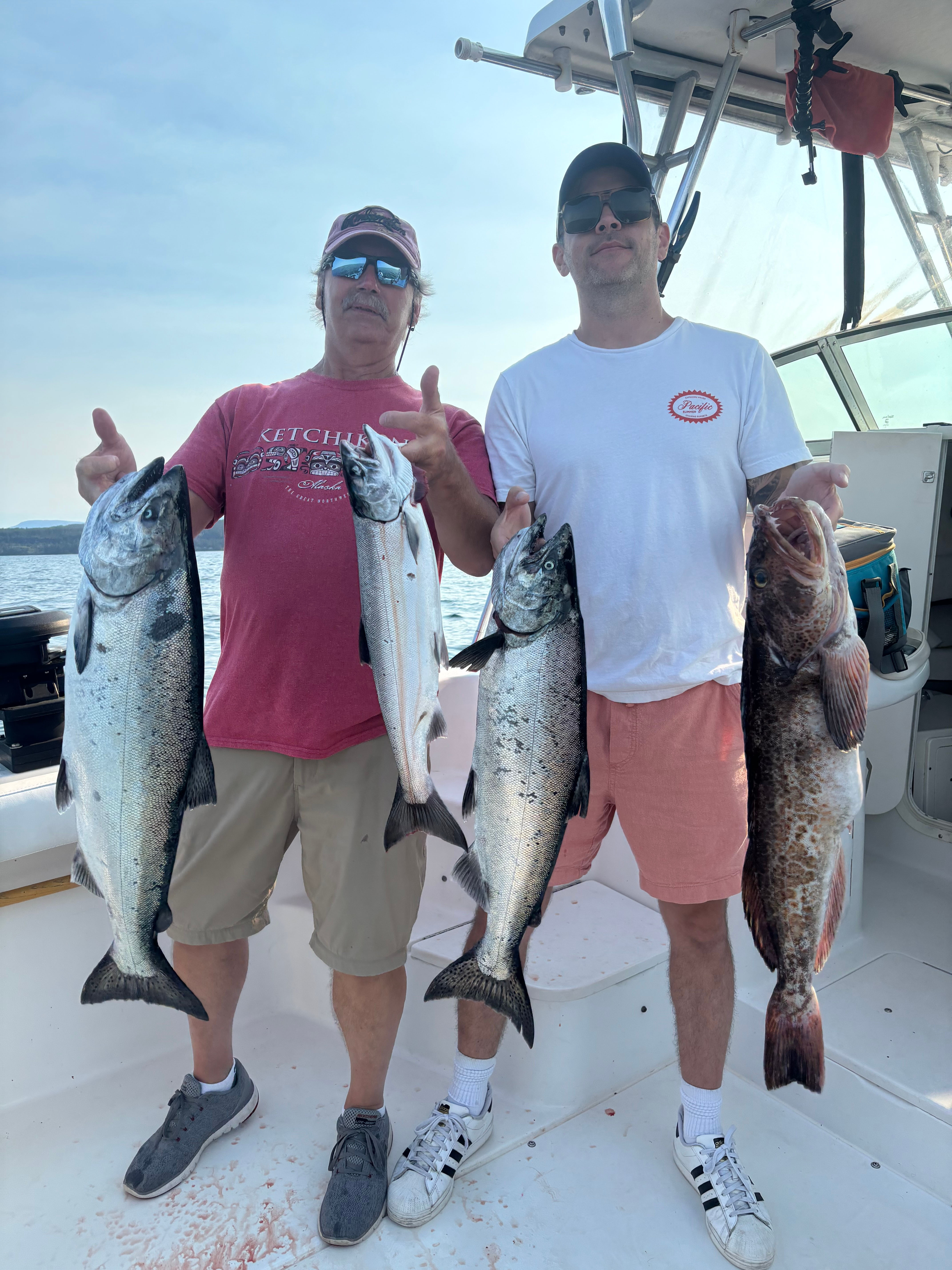 Two men on a boat holding four large fish they caught, with water and distant shore in the background.