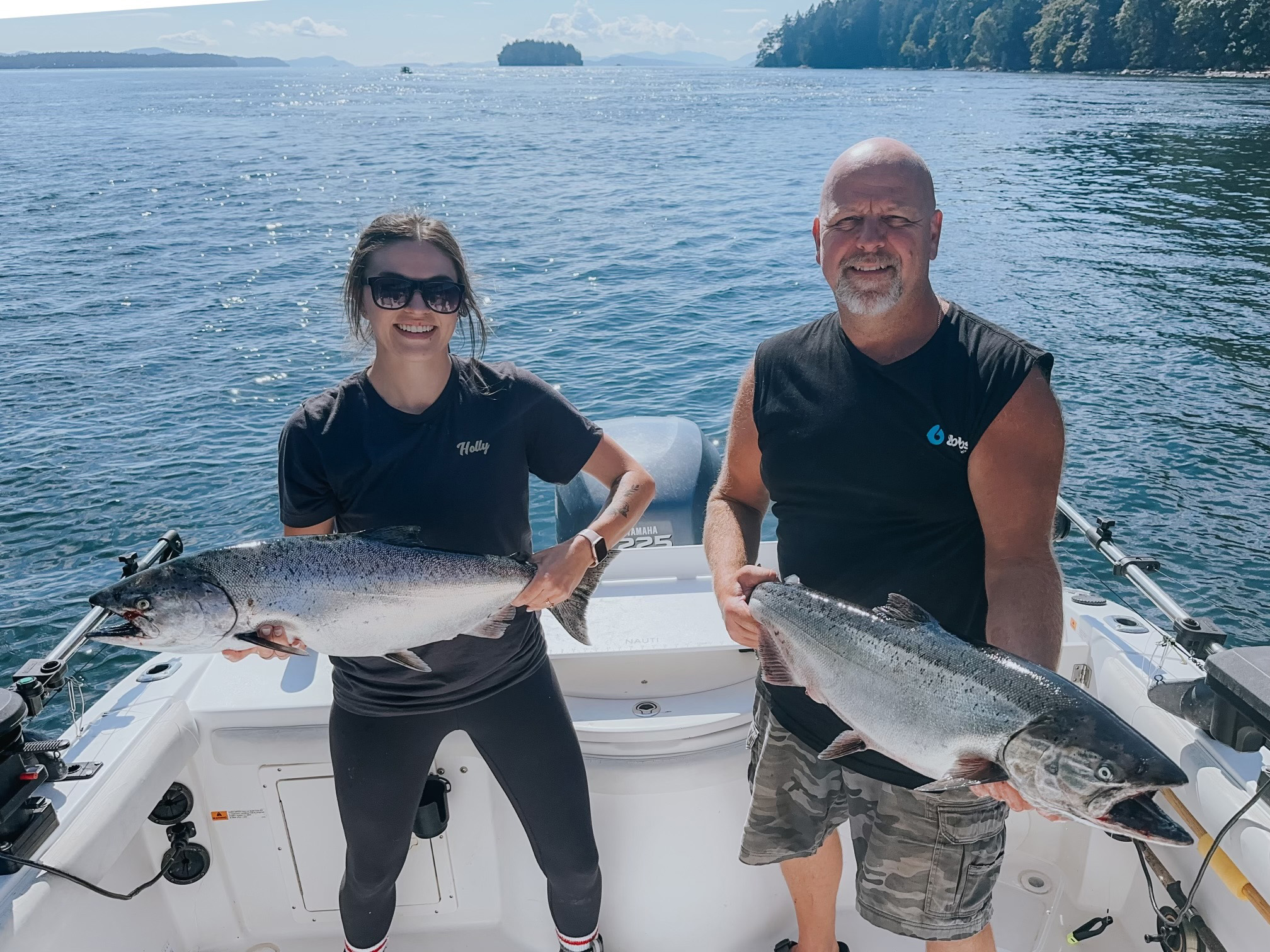 Two people standing on a boat holding large fish with a calm sea and forested shoreline in the background.