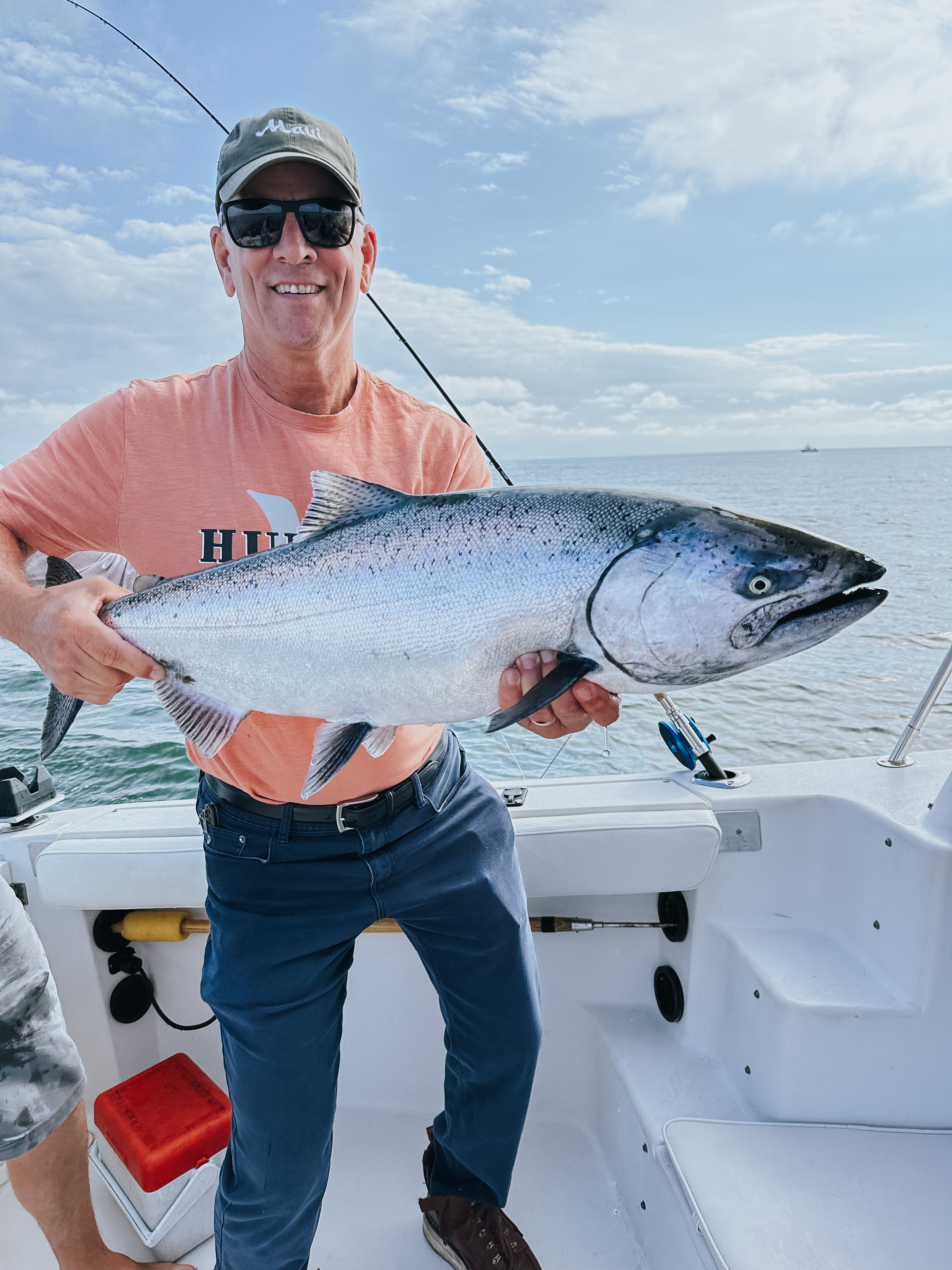 Man wearing sunglasses and a cap holding a large silver fish on a boat with ocean and cloudy sky in the background.