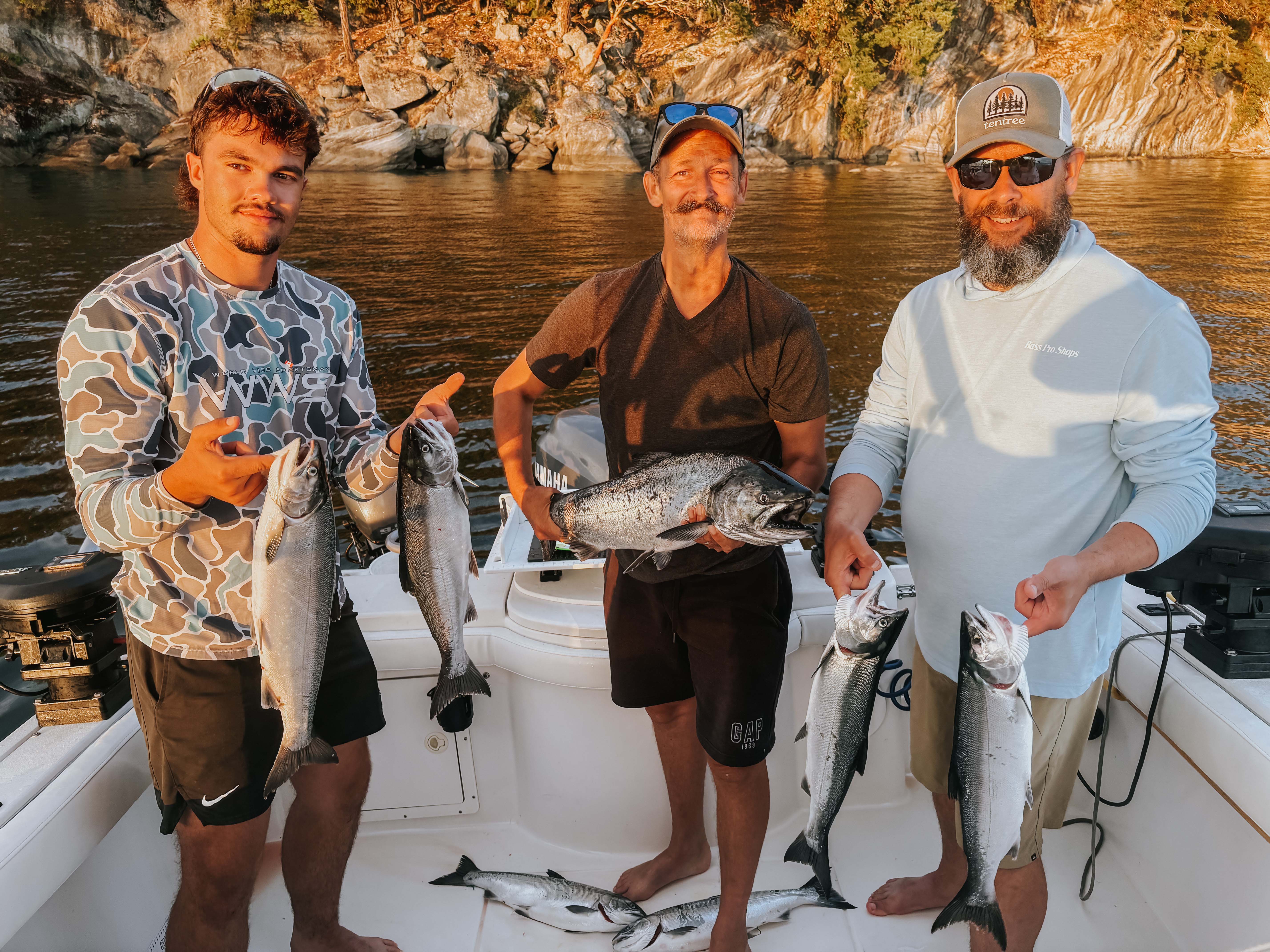 Three men on a boat holding freshly caught fish with a rocky shoreline and water in the background.