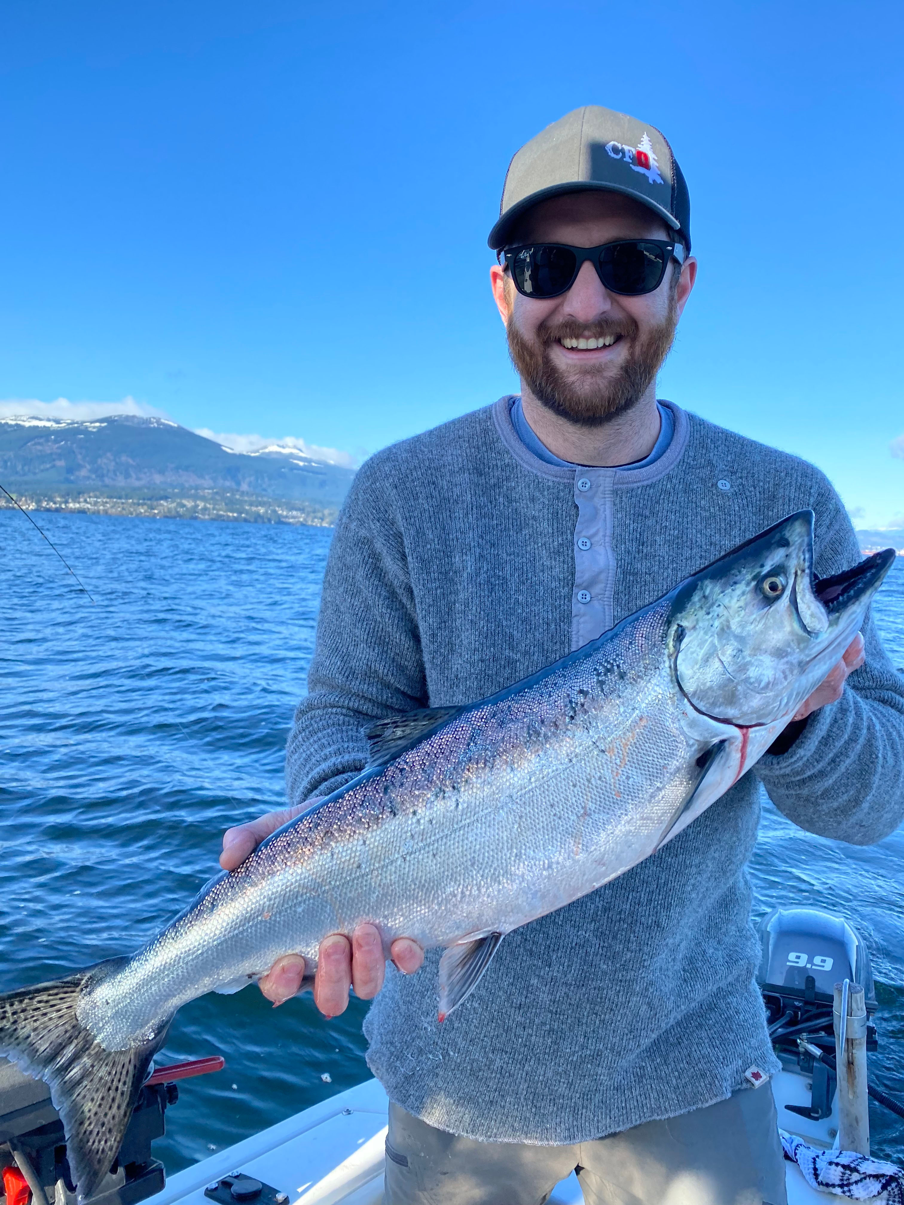 Smiling man wearing sunglasses and a cap holding a large silver fish on a boat with the ocean and mountains in the background.