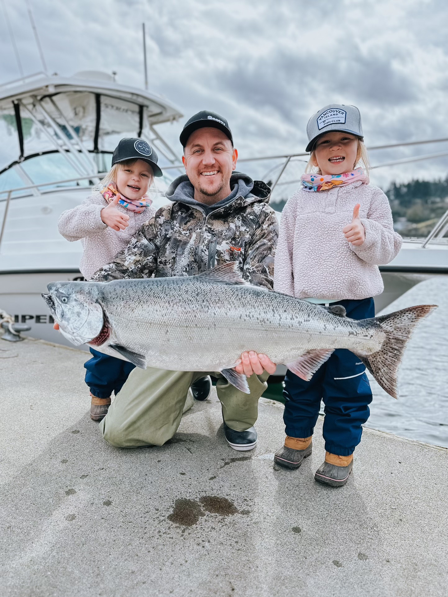 Man kneeling on dock holding a large silver fish with two young girls on either side giving thumbs up, with a boat and cloudy sky in the background.