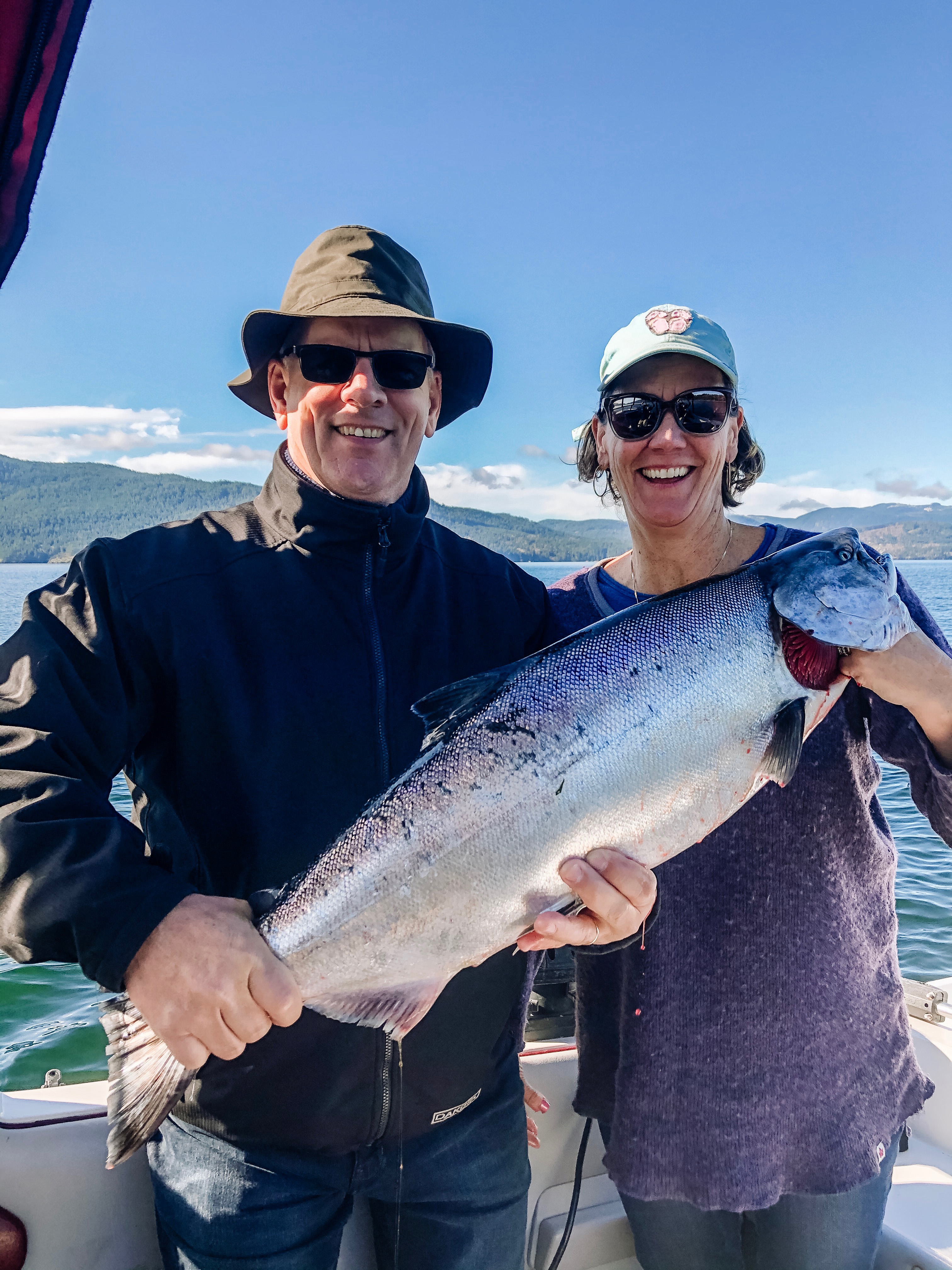 Two smiling people wearing sunglasses holding a large fish on a boat with water and hills in the background.
