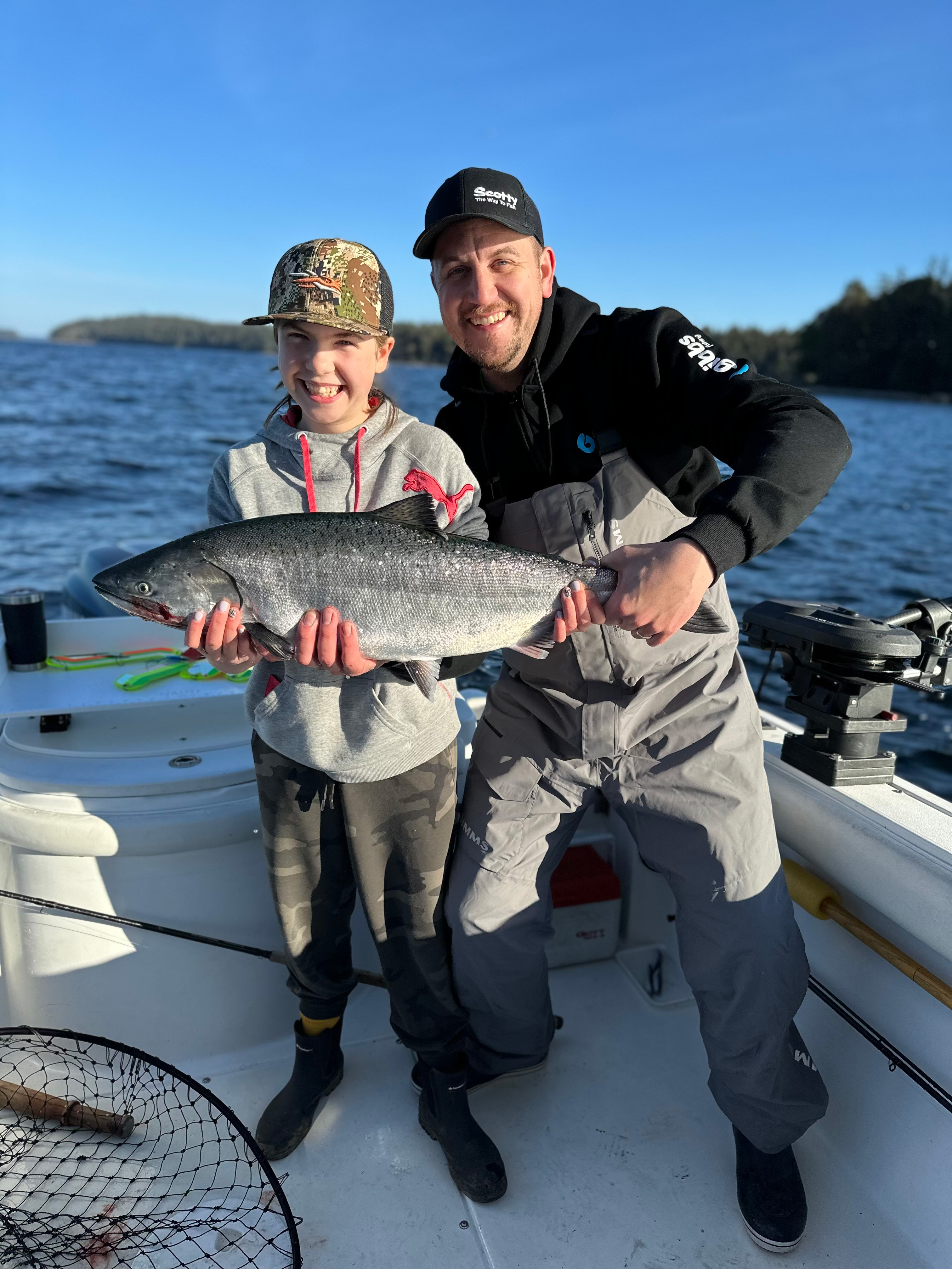 A smiling man and child holding a large fish together on a boat with water and trees in the background.