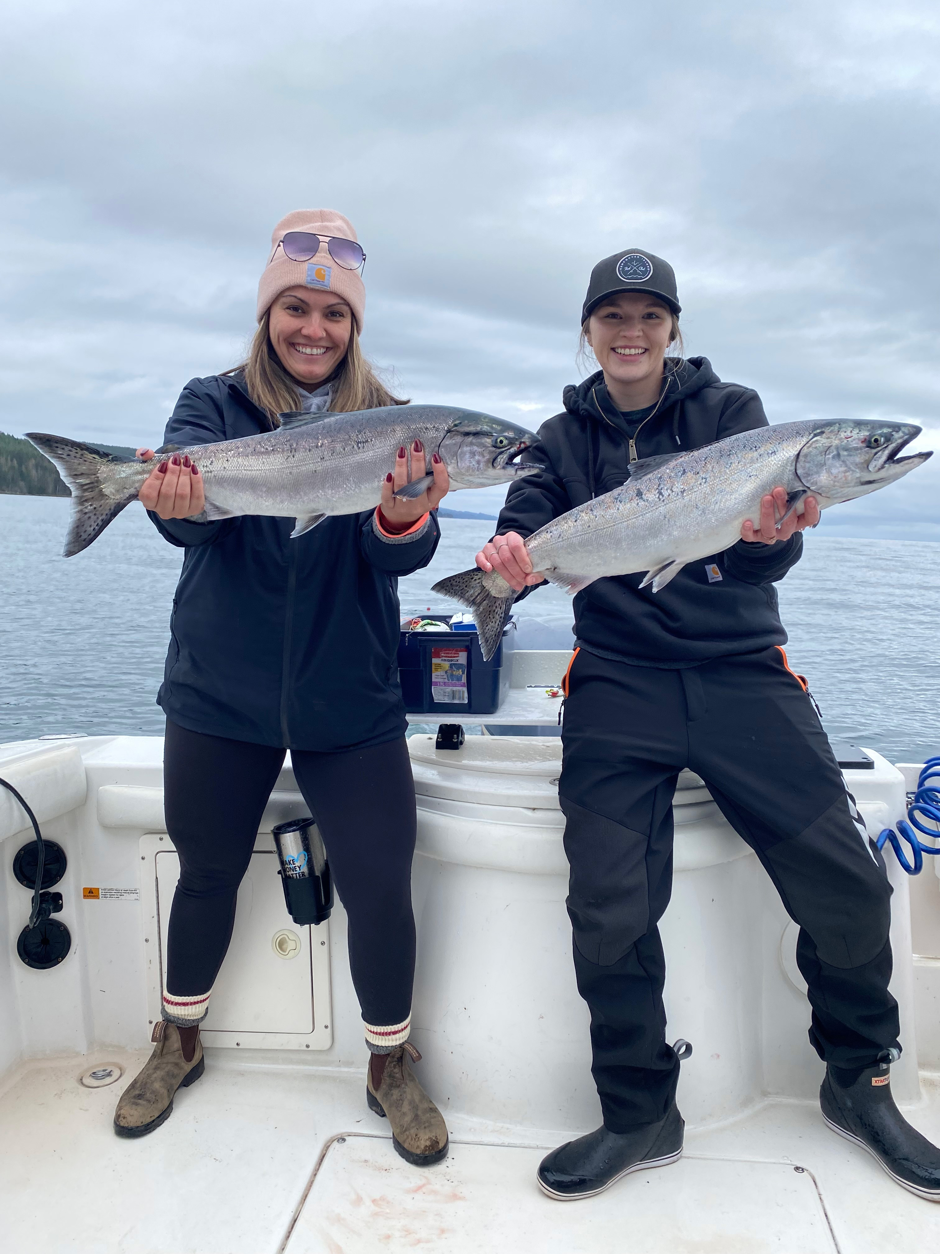 Two women on a boat holding large silver fish against a cloudy sky and ocean backdrop.