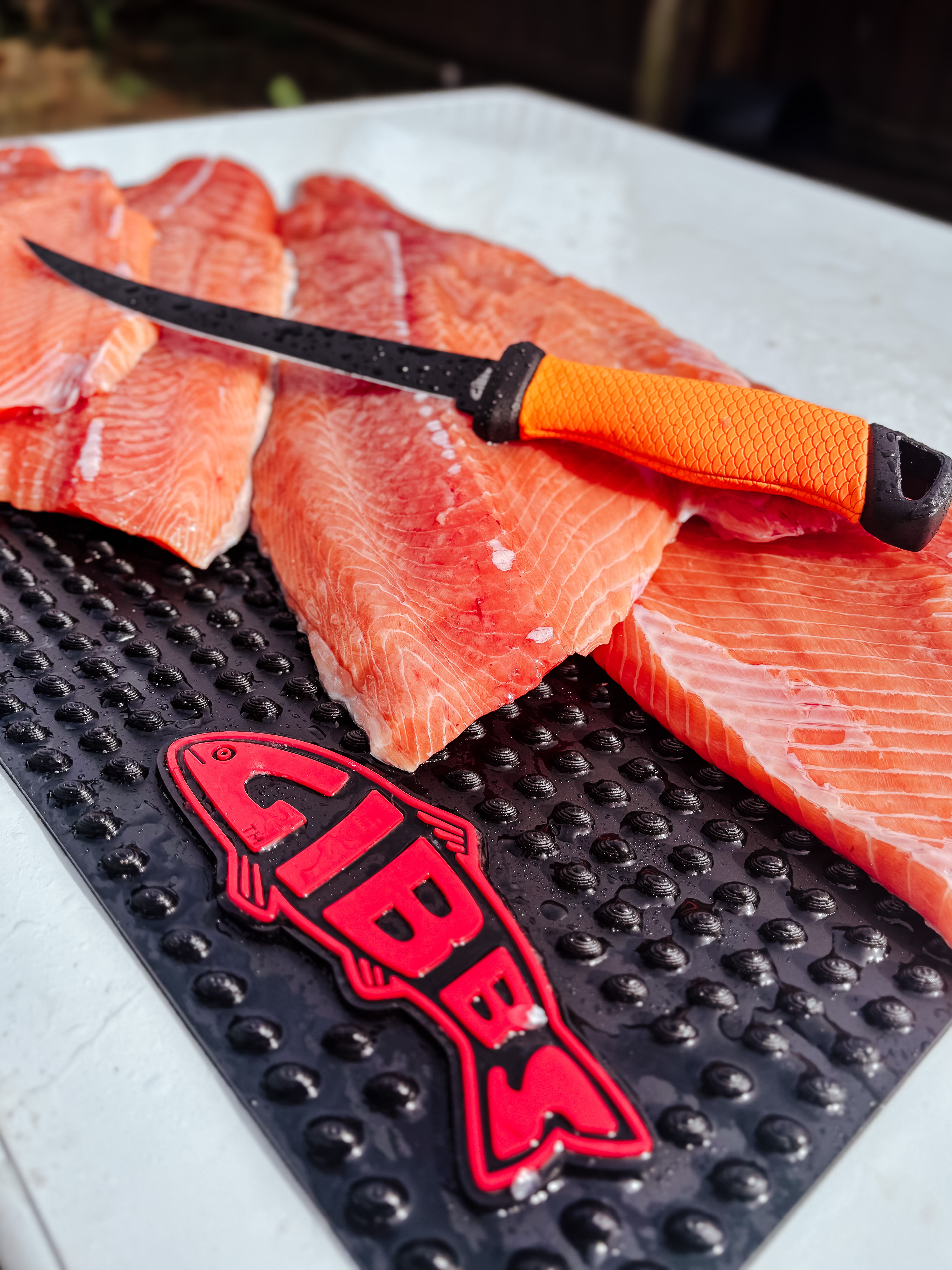 Fresh salmon fillets and a fillet knife with an orange handle on a black CIBBS-branded cutting board.