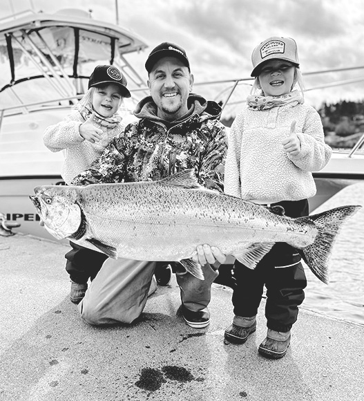 Man kneeling on a dock holding a large fish, flanked by two children giving thumbs up, with a boat in the background.