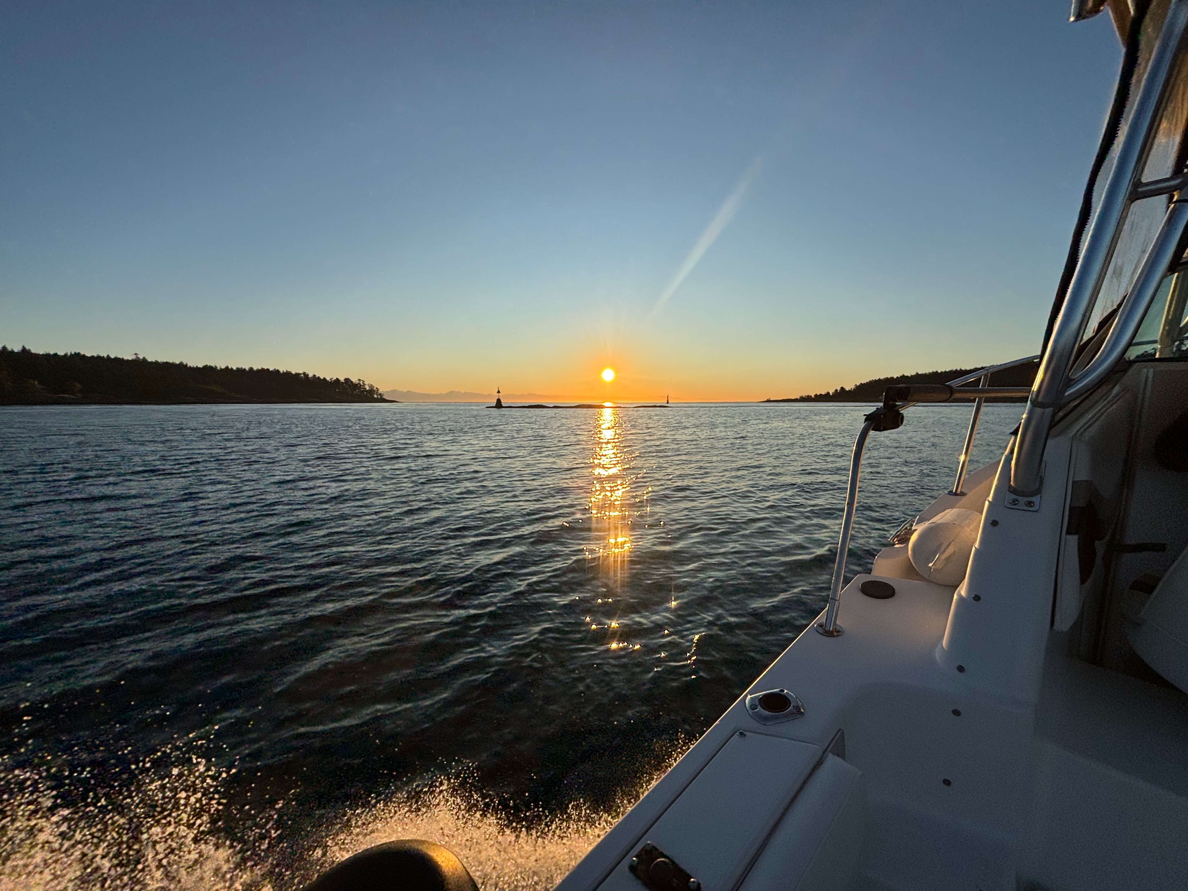 Sun setting over calm ocean waters viewed from the side of a white boat with land silhouettes on the horizon.