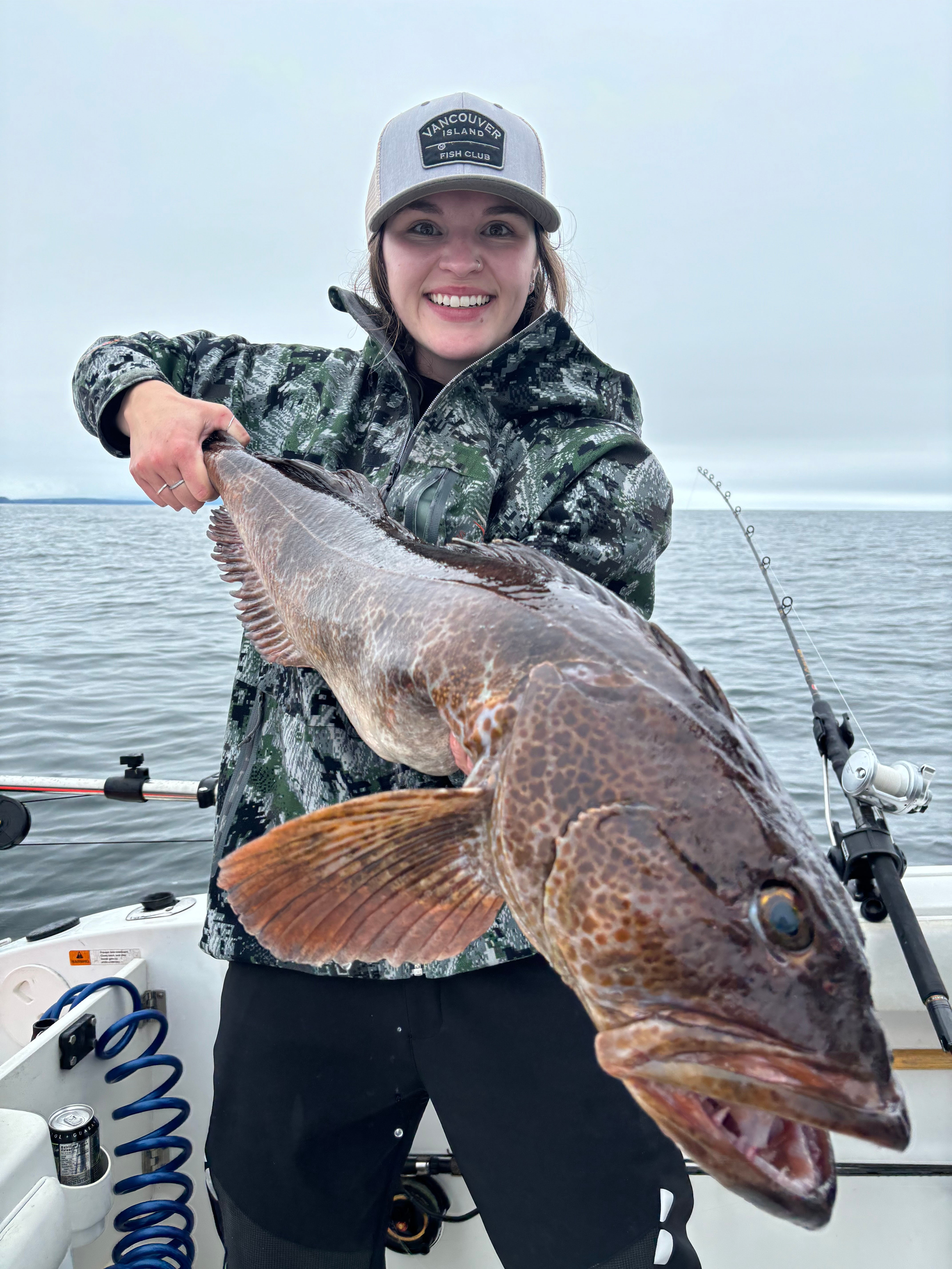 Smiling woman in a camouflage jacket and cap holding a large lingcod fish on a boat with ocean in the background.