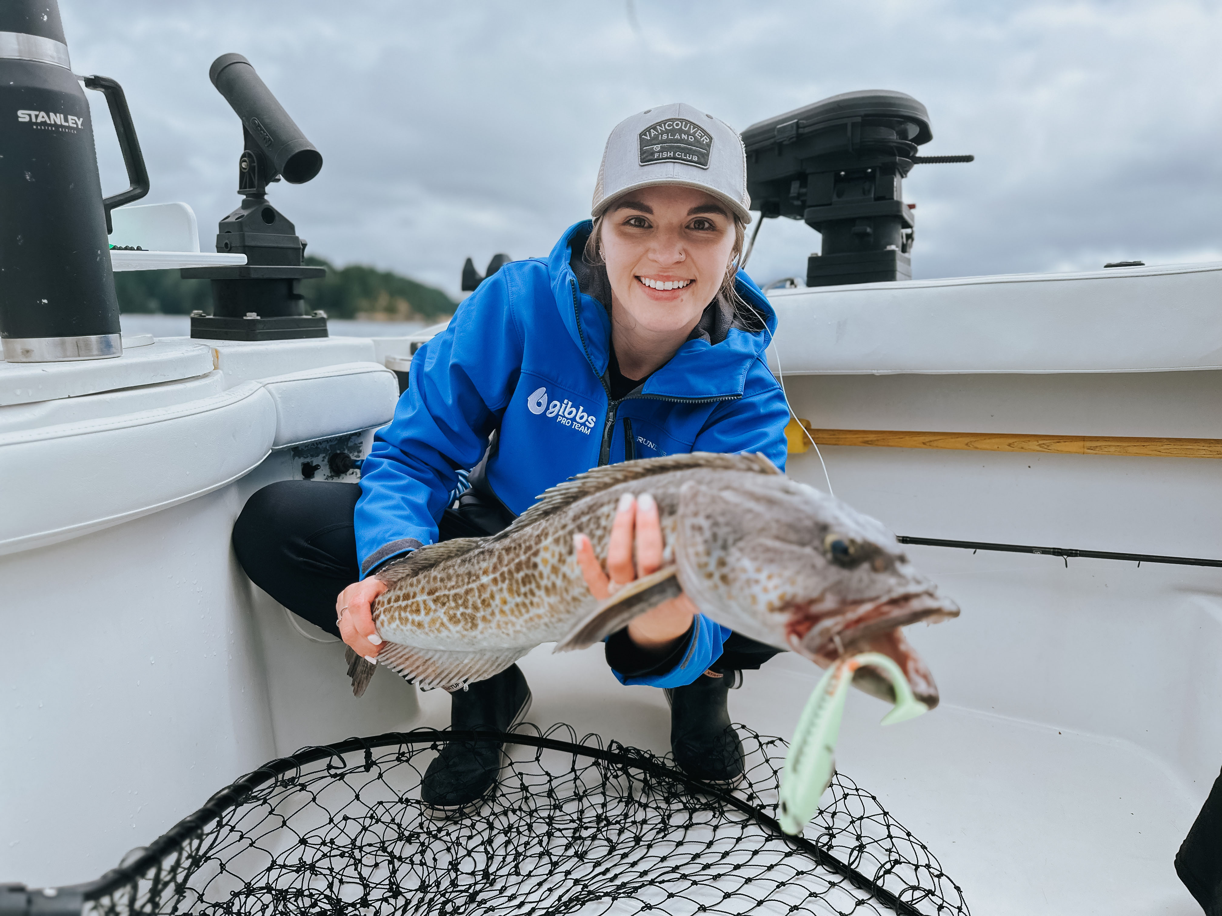 Woman in blue jacket and cap smiling while holding a large lingcod fish on a boat.