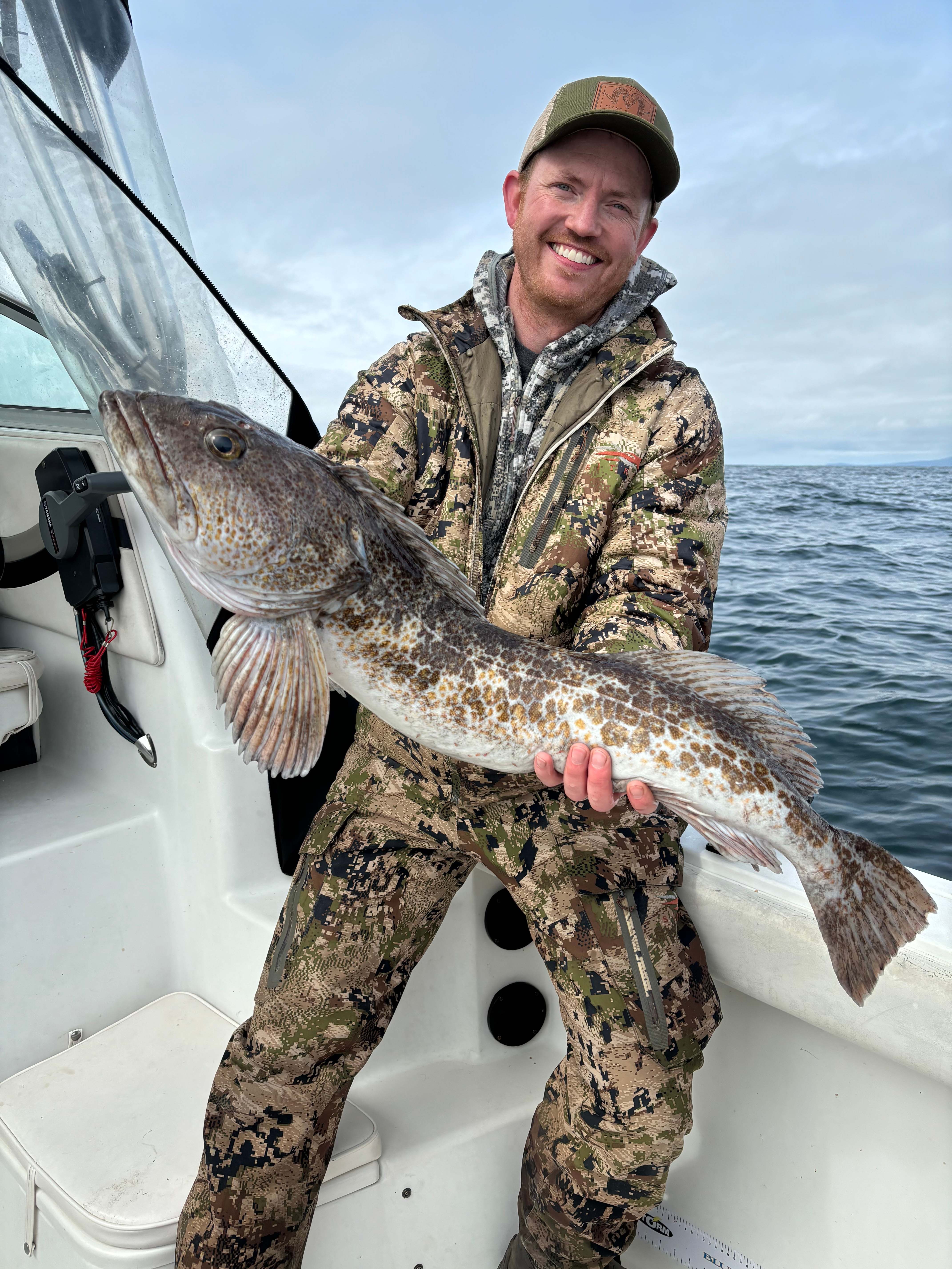 Smiling man in camouflage jacket and hat holding a large lingcod fish on a boat with ocean in the background.