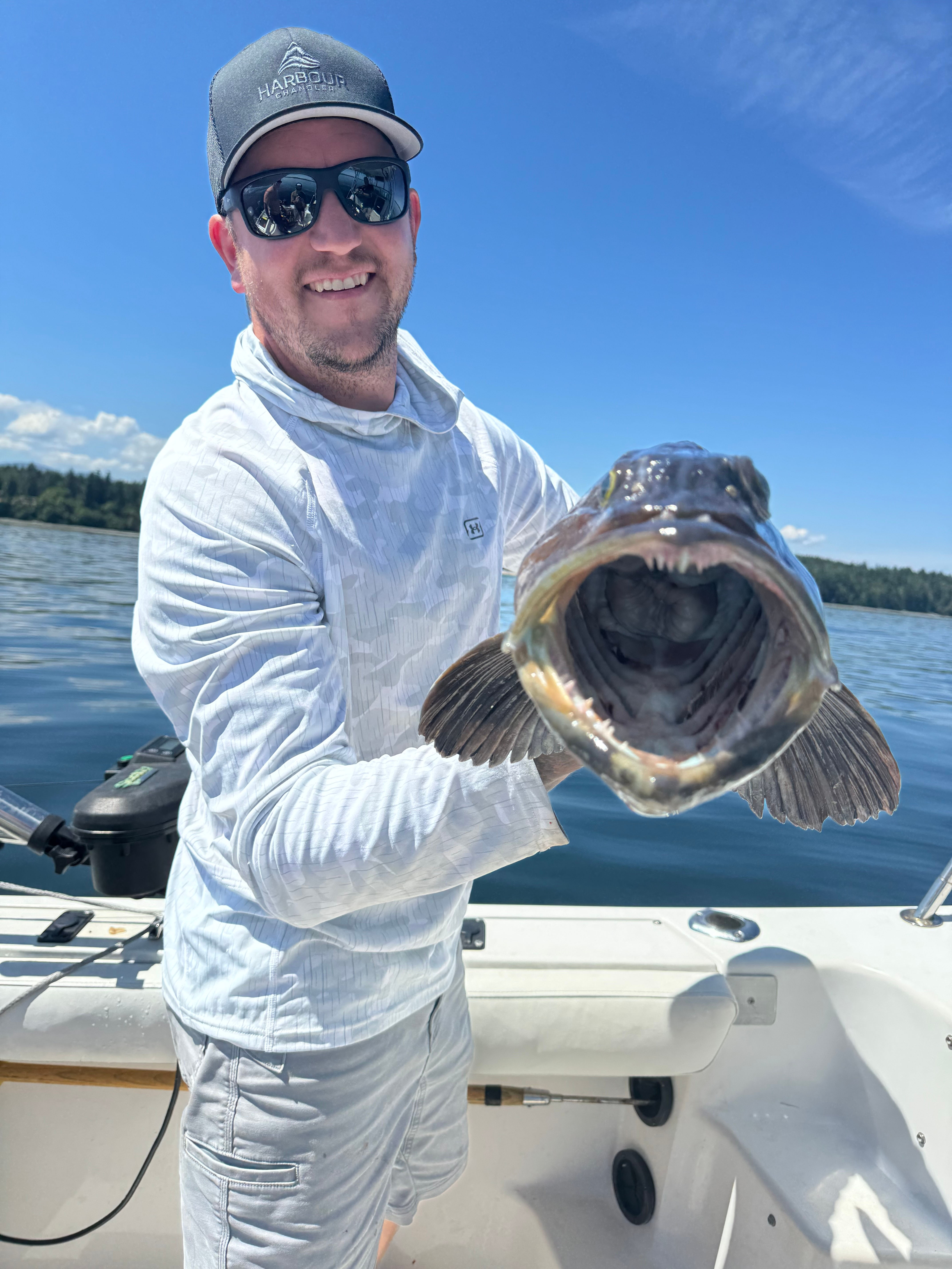 Man wearing sunglasses and cap smiling while holding a large lingcod with an open mouth on a boat