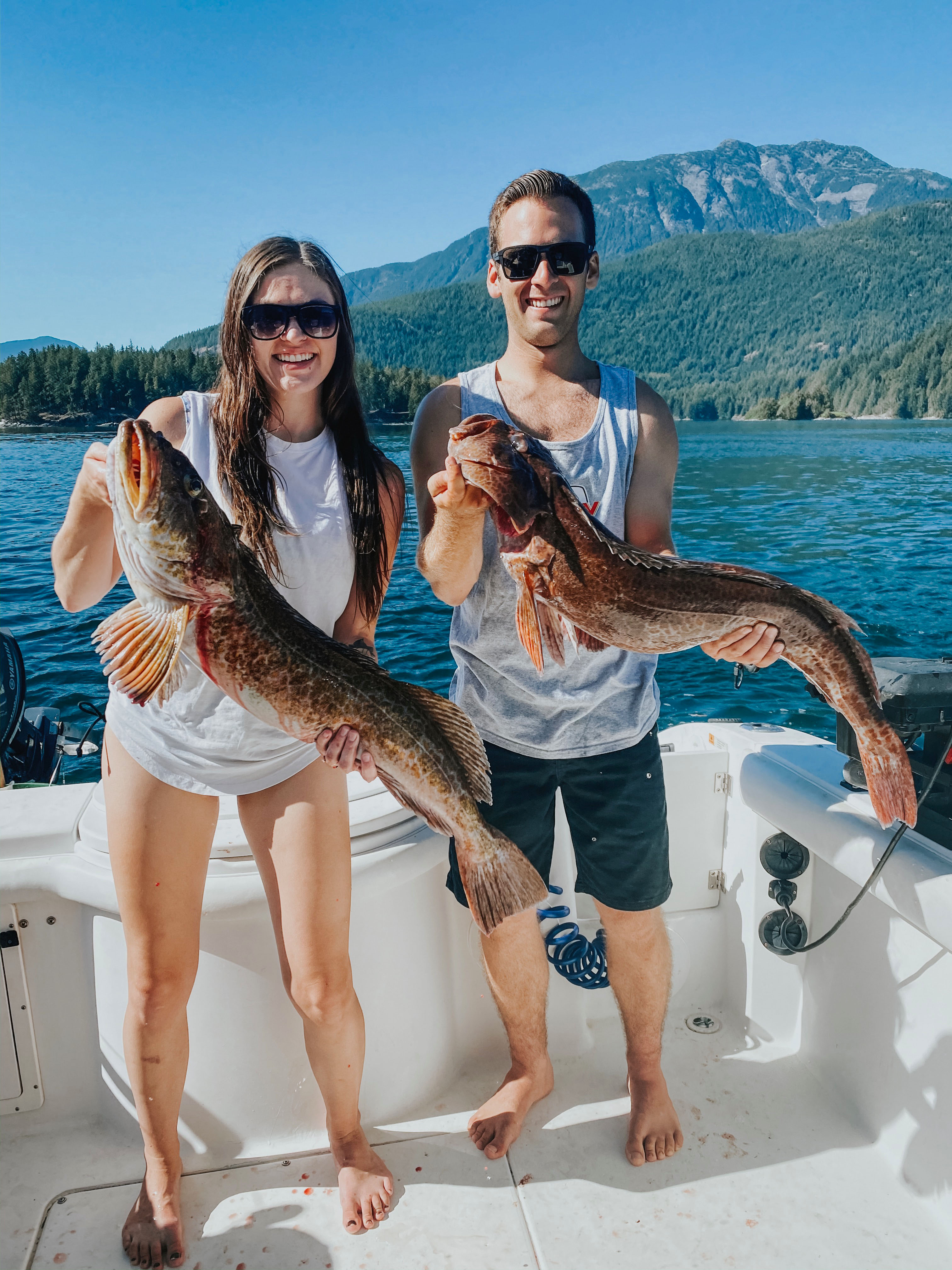 Smiling man and woman on a boat holding large lingcod fish with mountains and forest in the background.