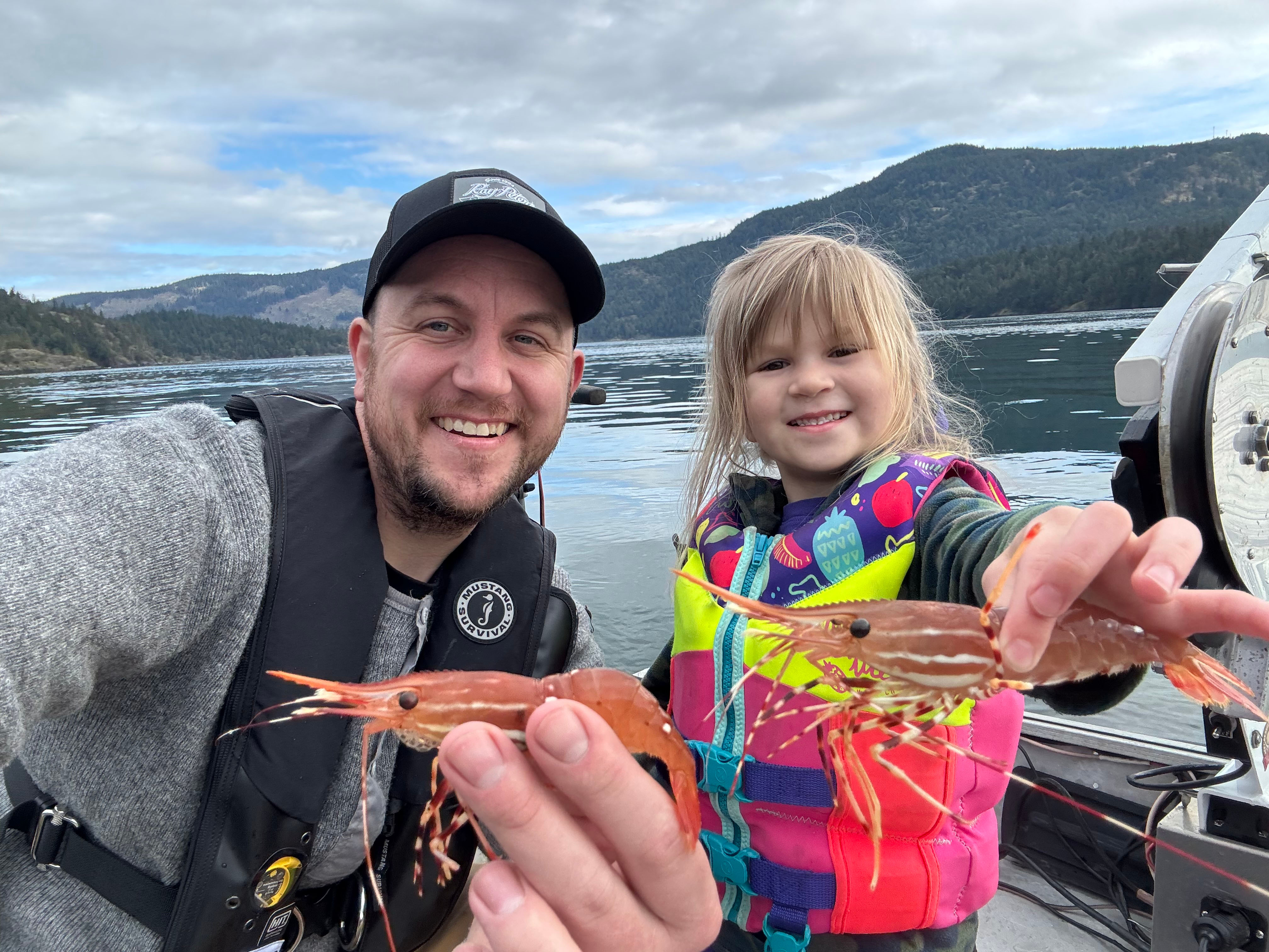Smiling man and young girl on a boat holding up two large prawns with water and forested hills in the background.