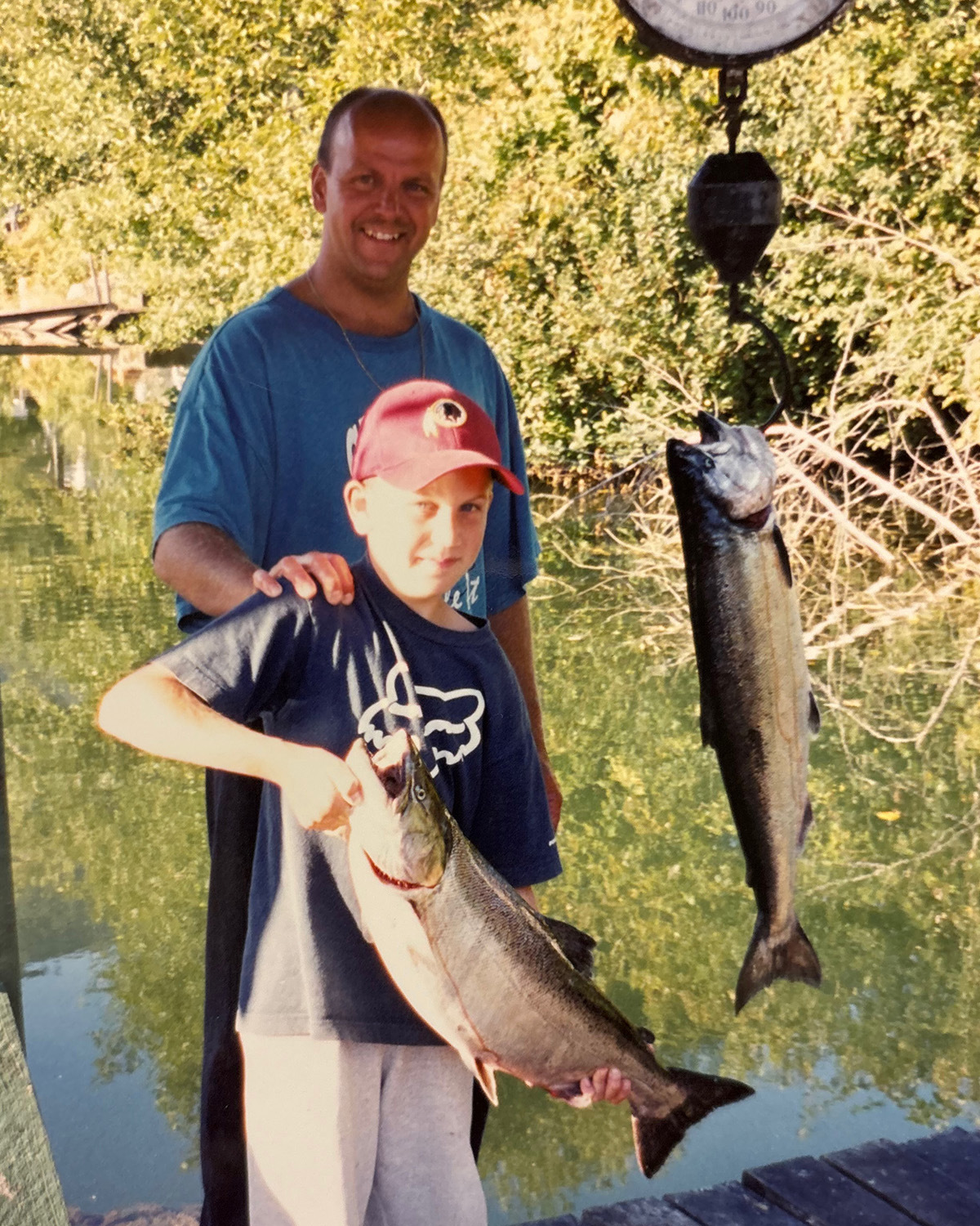 A smiling man stands behind a boy holding a large fish, with another fish hanging on a scale by a riverbank.