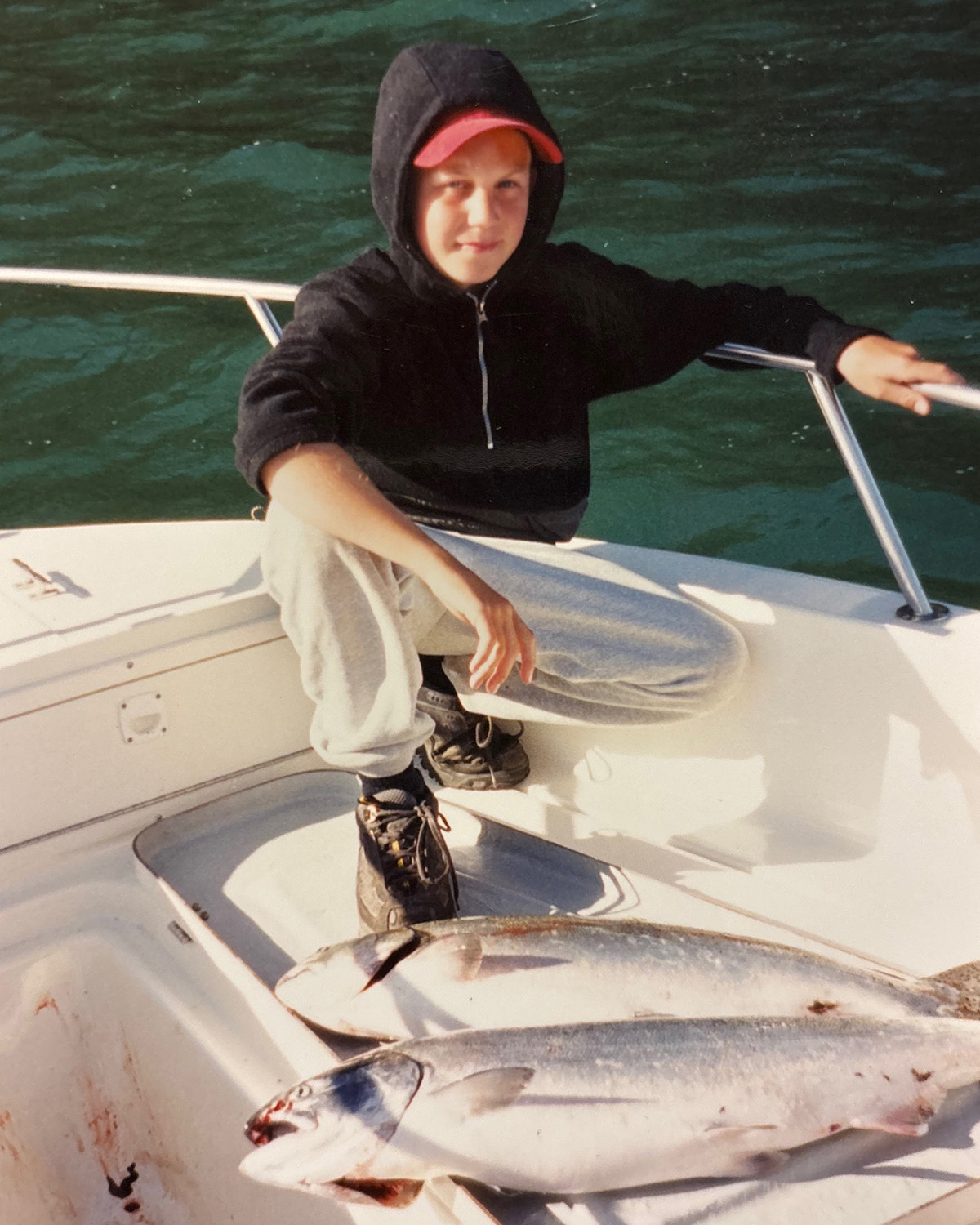 Young person wearing a black hoodie and red cap crouching on a boat next to two large caught fish.