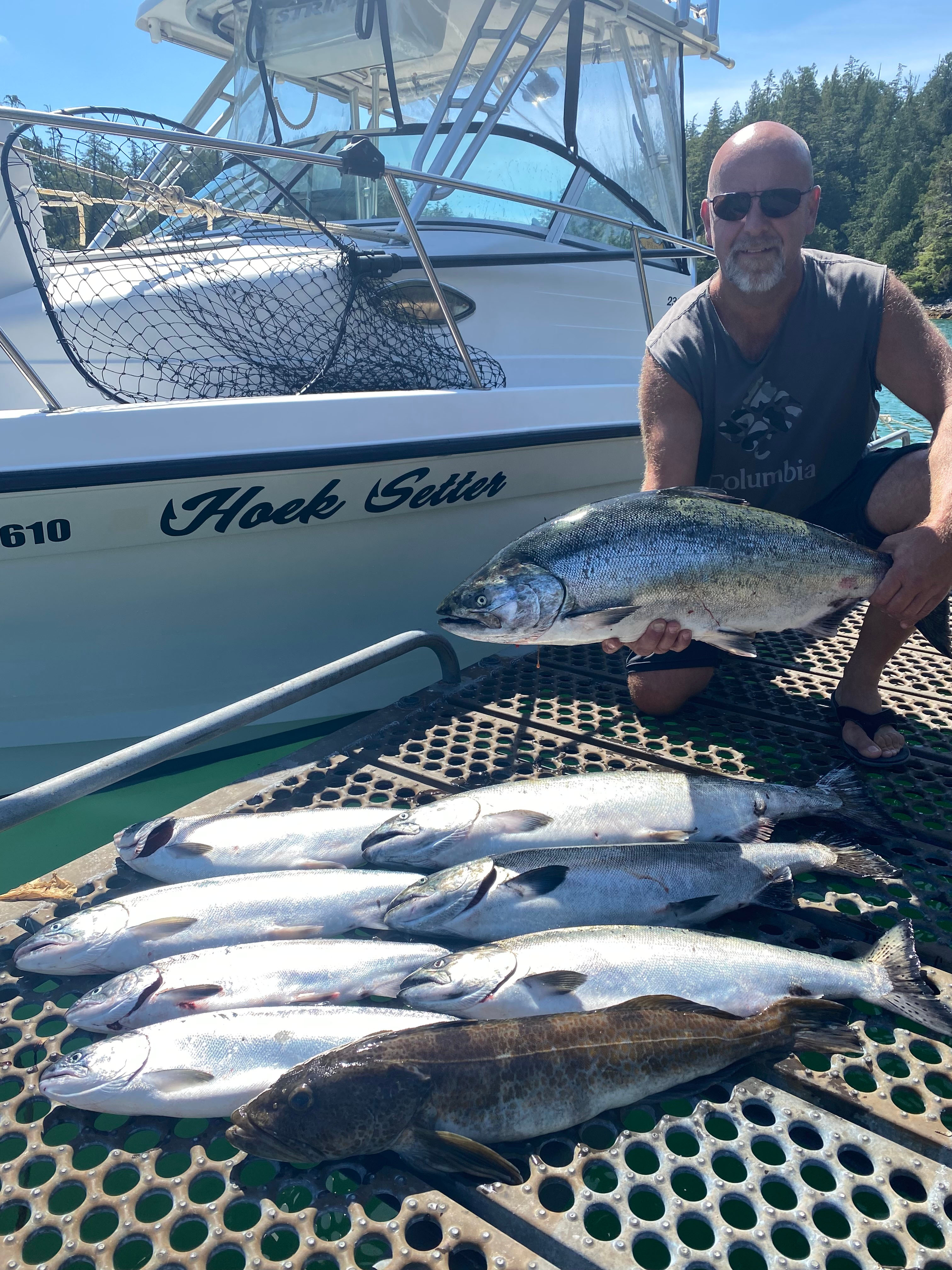 Man wearing sunglasses holding a large fish beside a boat named 'Hoek Setter' with six other fish laid out on a perforated dock.