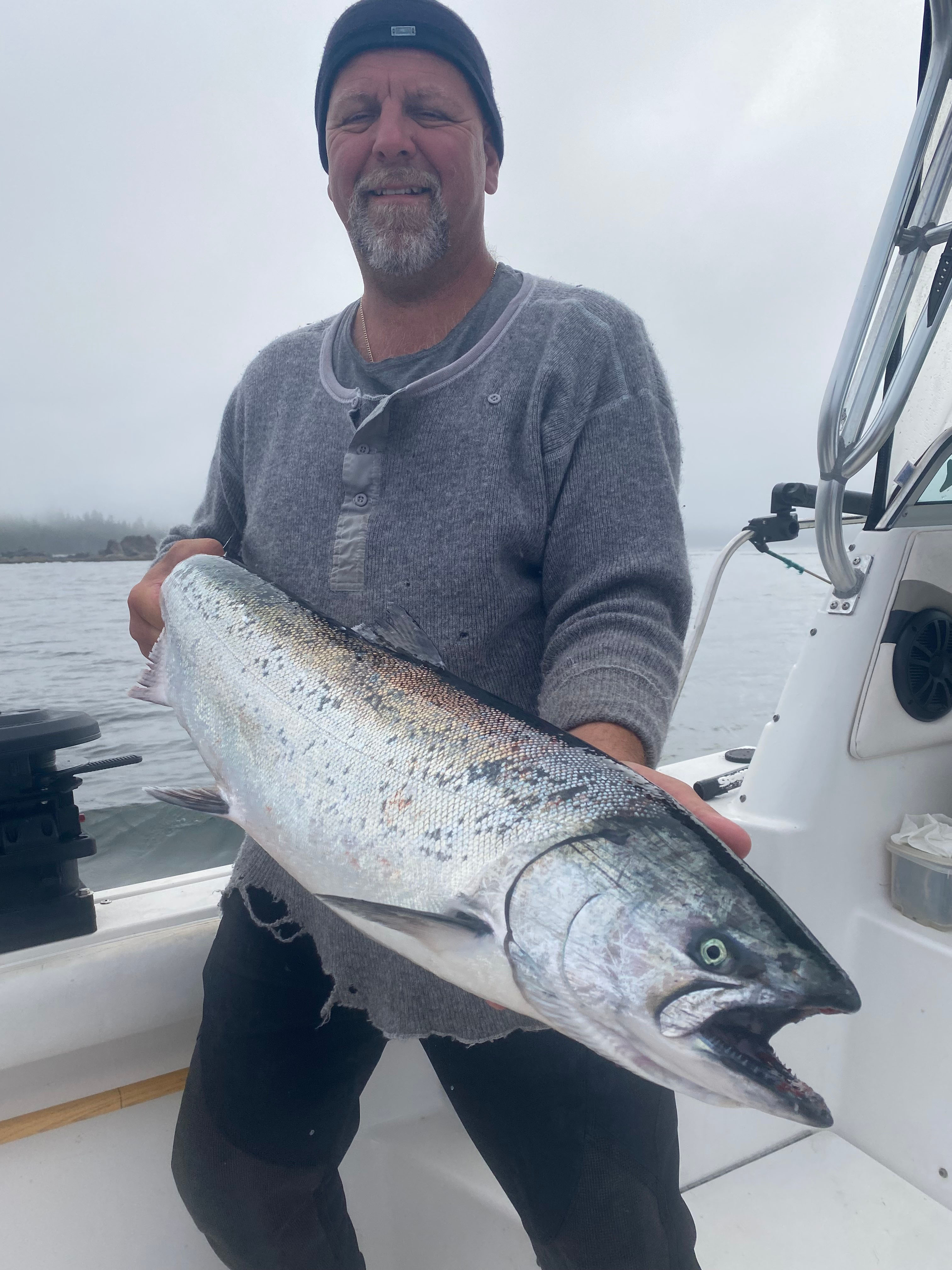 Smiling man wearing a beanie and gray sweater holding a large silver fish on a boat near a foggy shoreline.
