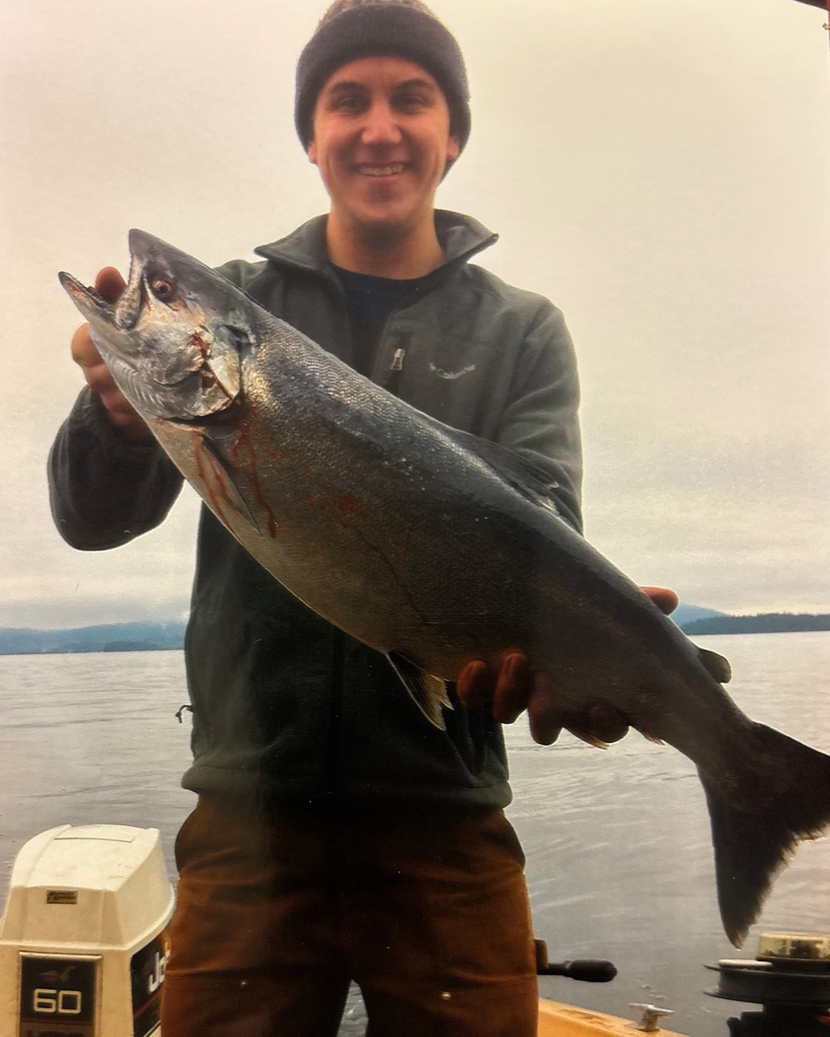 Smiling man in a beanie holding a large fish on a boat with water and cloudy sky in the background.