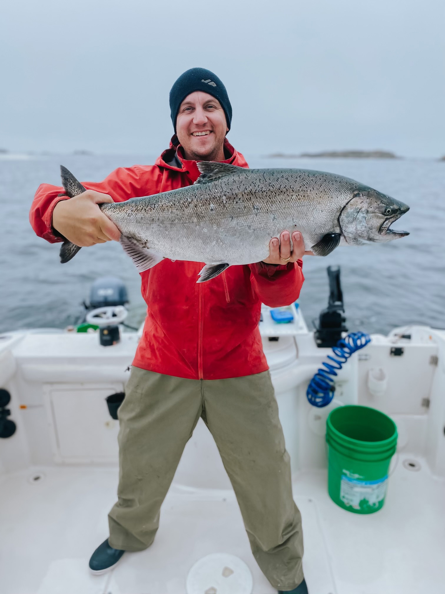 Smiling man in red jacket and black beanie holding a large fish on a boat with water in the background.
