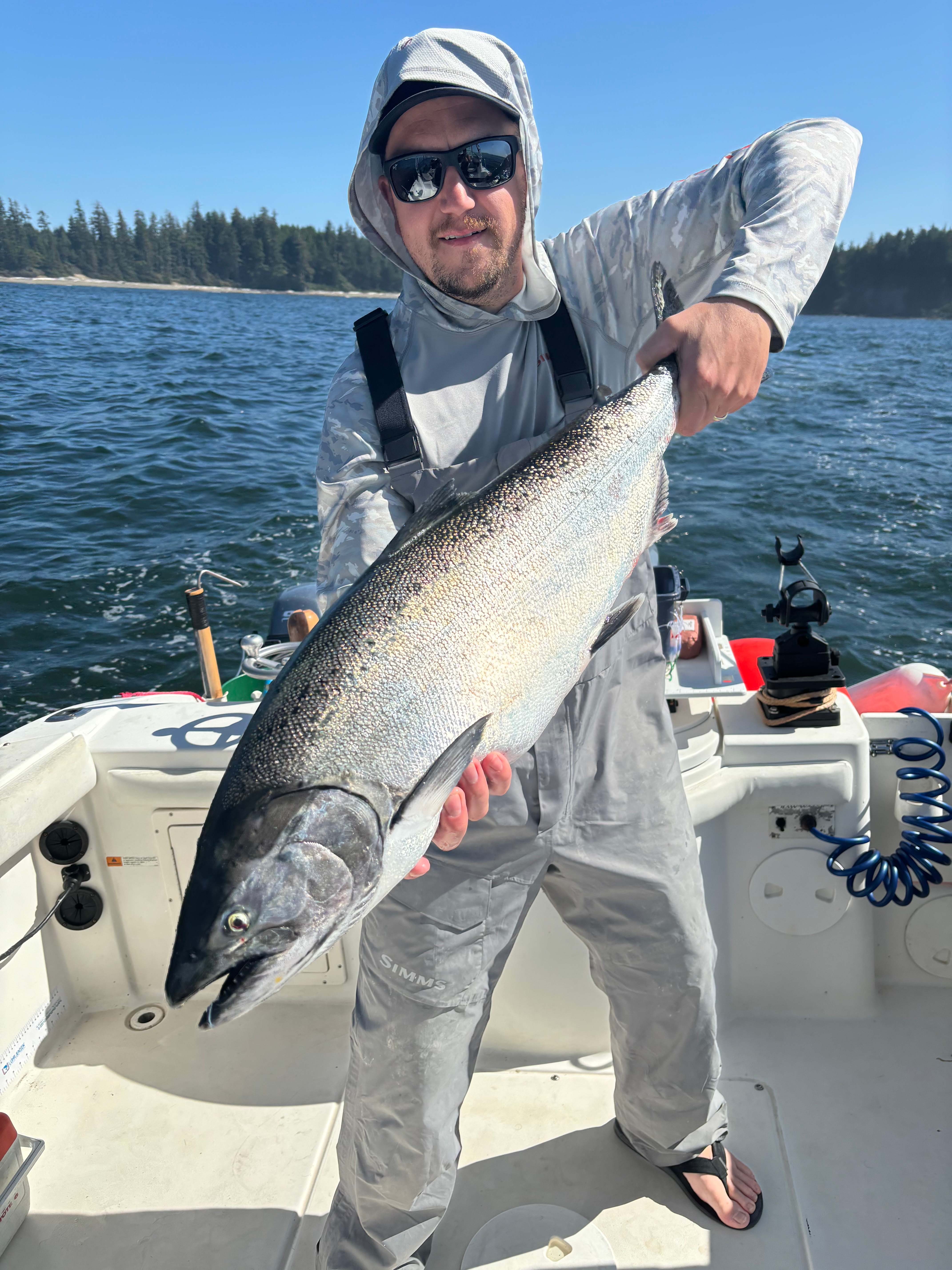 Man wearing sunglasses and gray fishing gear holding a large silver fish on a boat with ocean and forest in background.