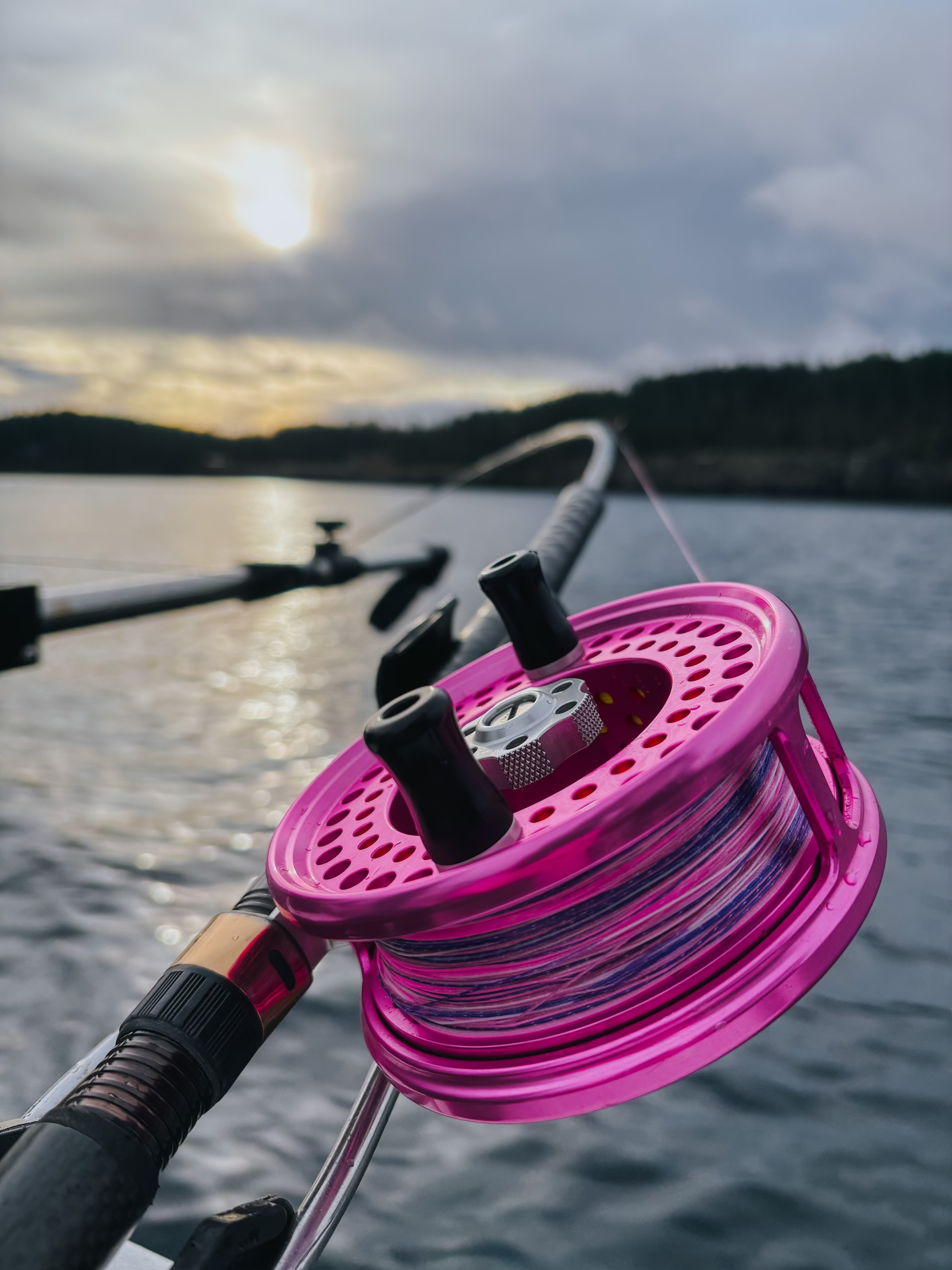 Close-up of a pink fishing reel attached to a fishing rod over the ocean at sunset.