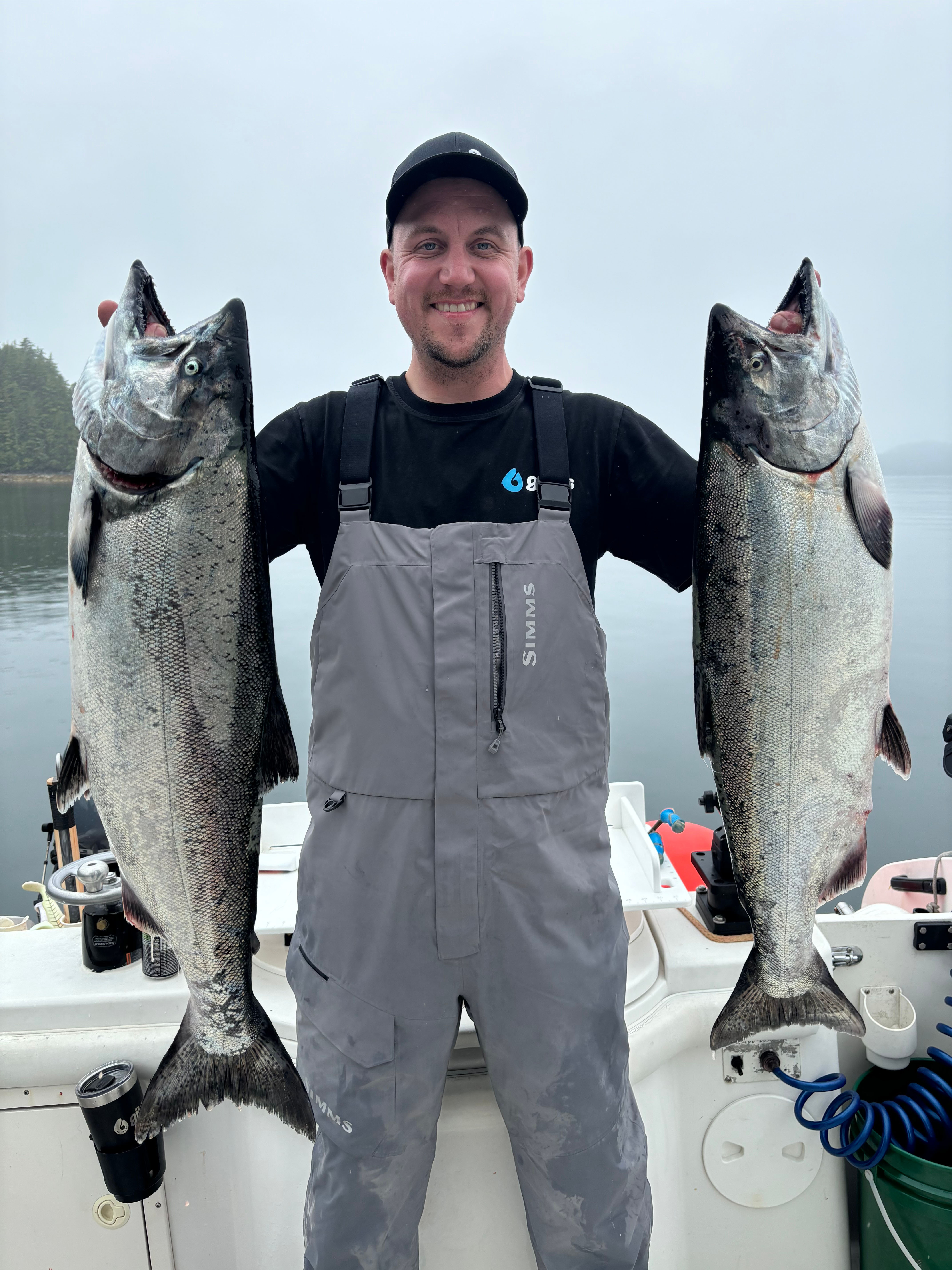 Smiling man in gray overalls holding two large caught fish on a boat with misty water and trees in the background.
