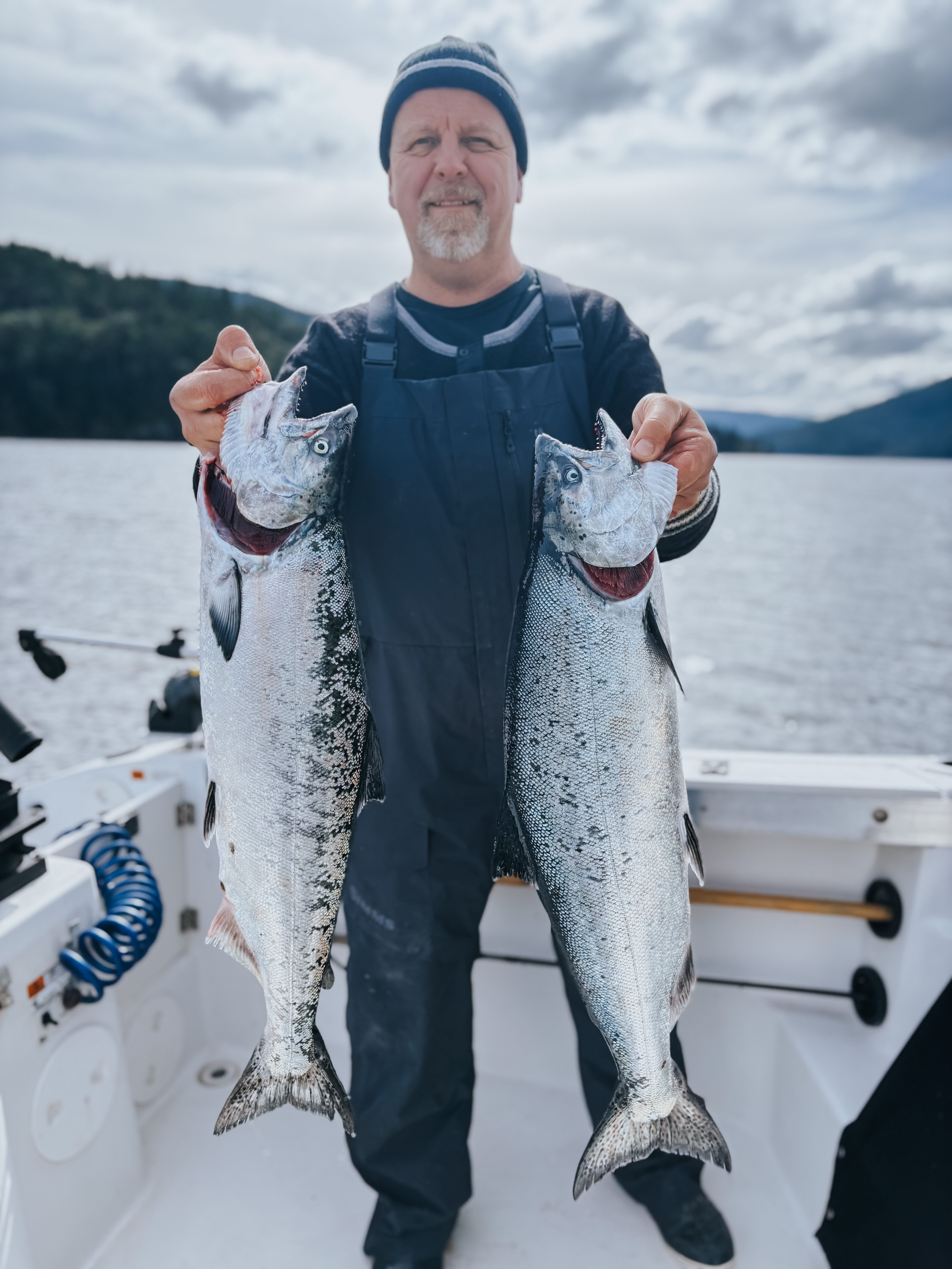 Man on a boat holding two large freshly caught silver fish with the ocean and forested hills in the background.