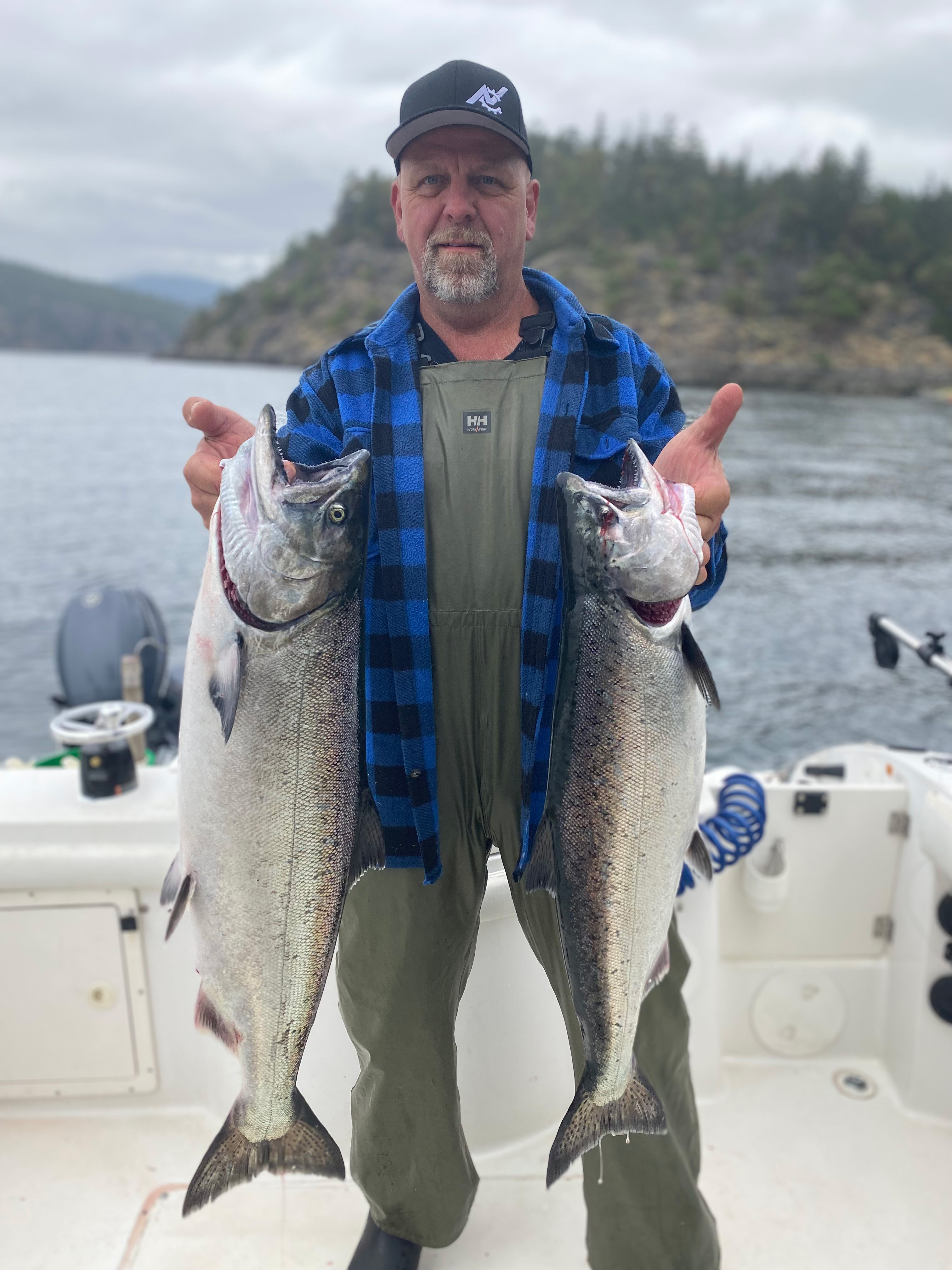 Man wearing green waders, blue plaid jacket, and black cap holding two large fish on a boat with water and forested hills in the background.