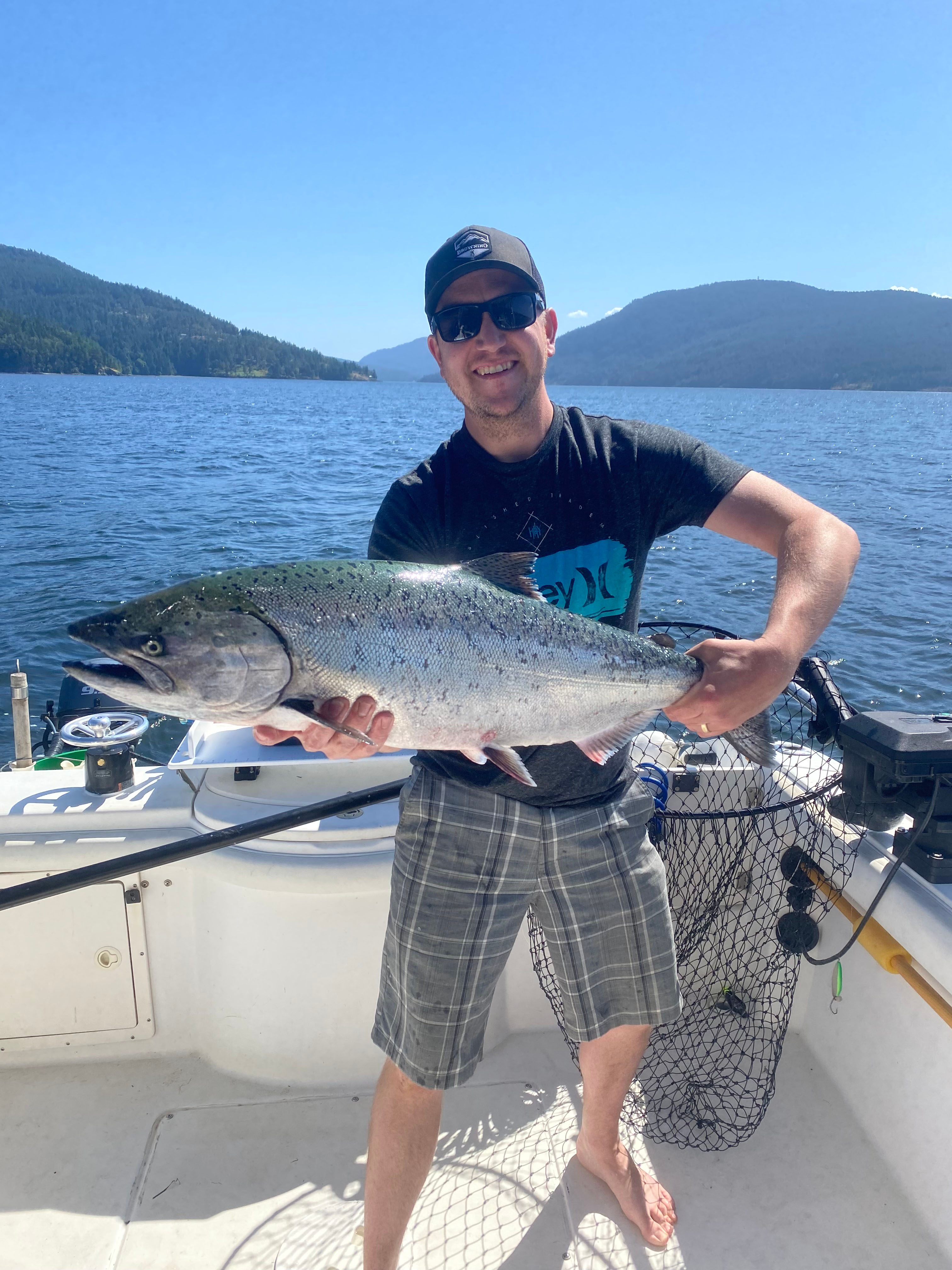 Man wearing sunglasses and a cap holding a large fish on a boat with water and forested hills in the background.