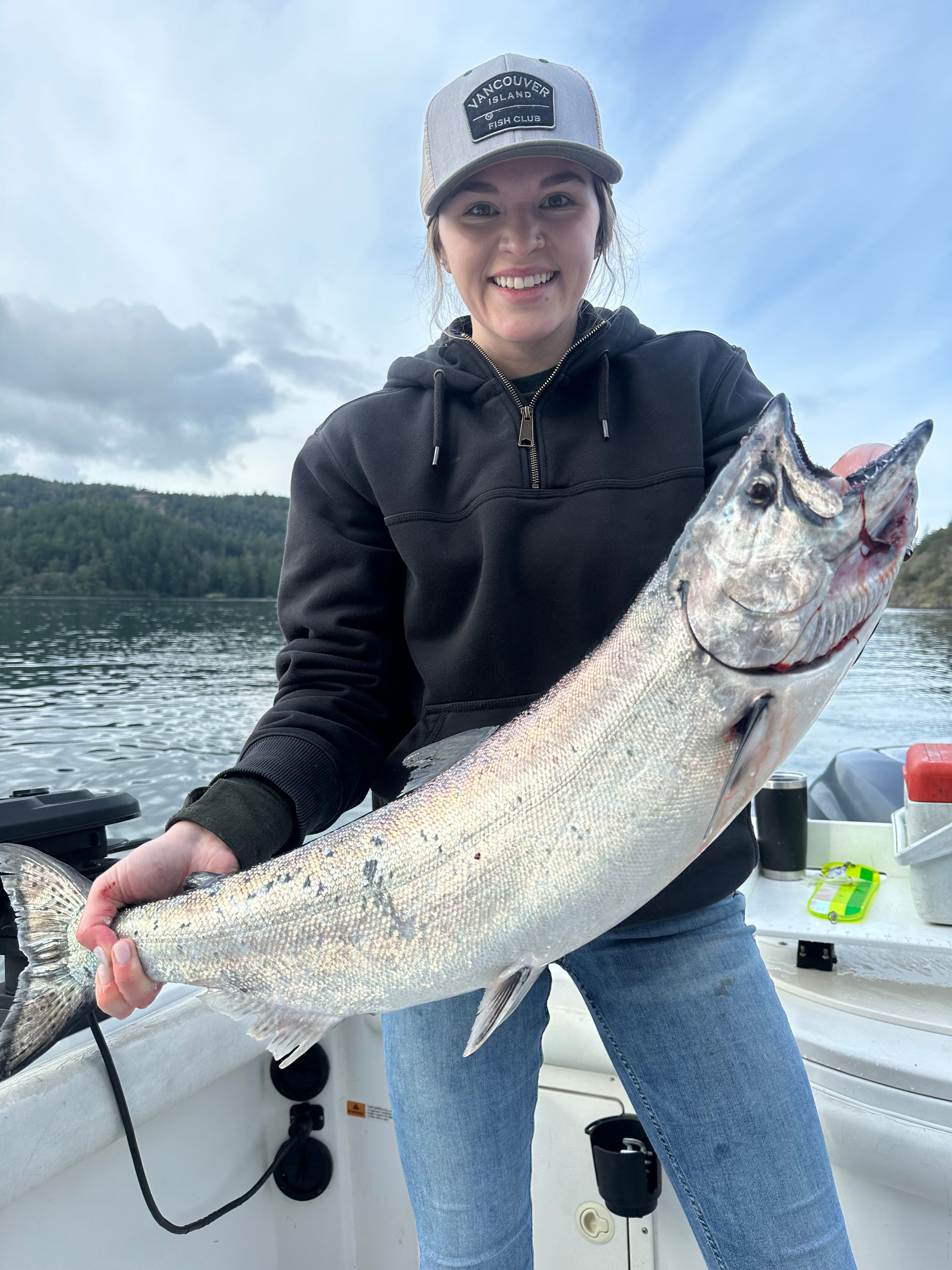 Smiling woman wearing a Vancouver Island Fish Club cap holding a large silver fish on a boat with water and forested hills in the background.