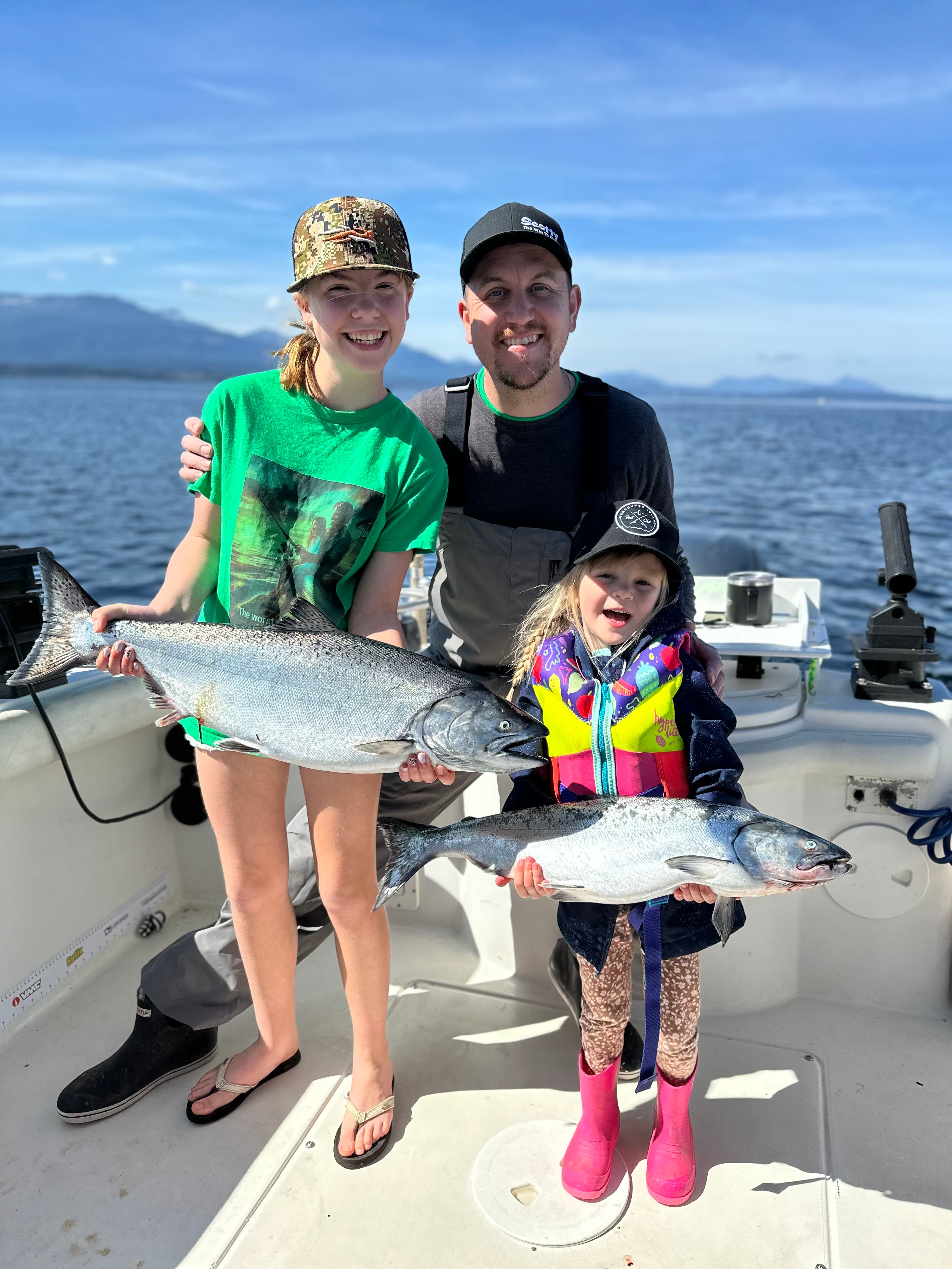Three people on a boat holding large silver fish, with water and mountains visible in the background on a sunny day.