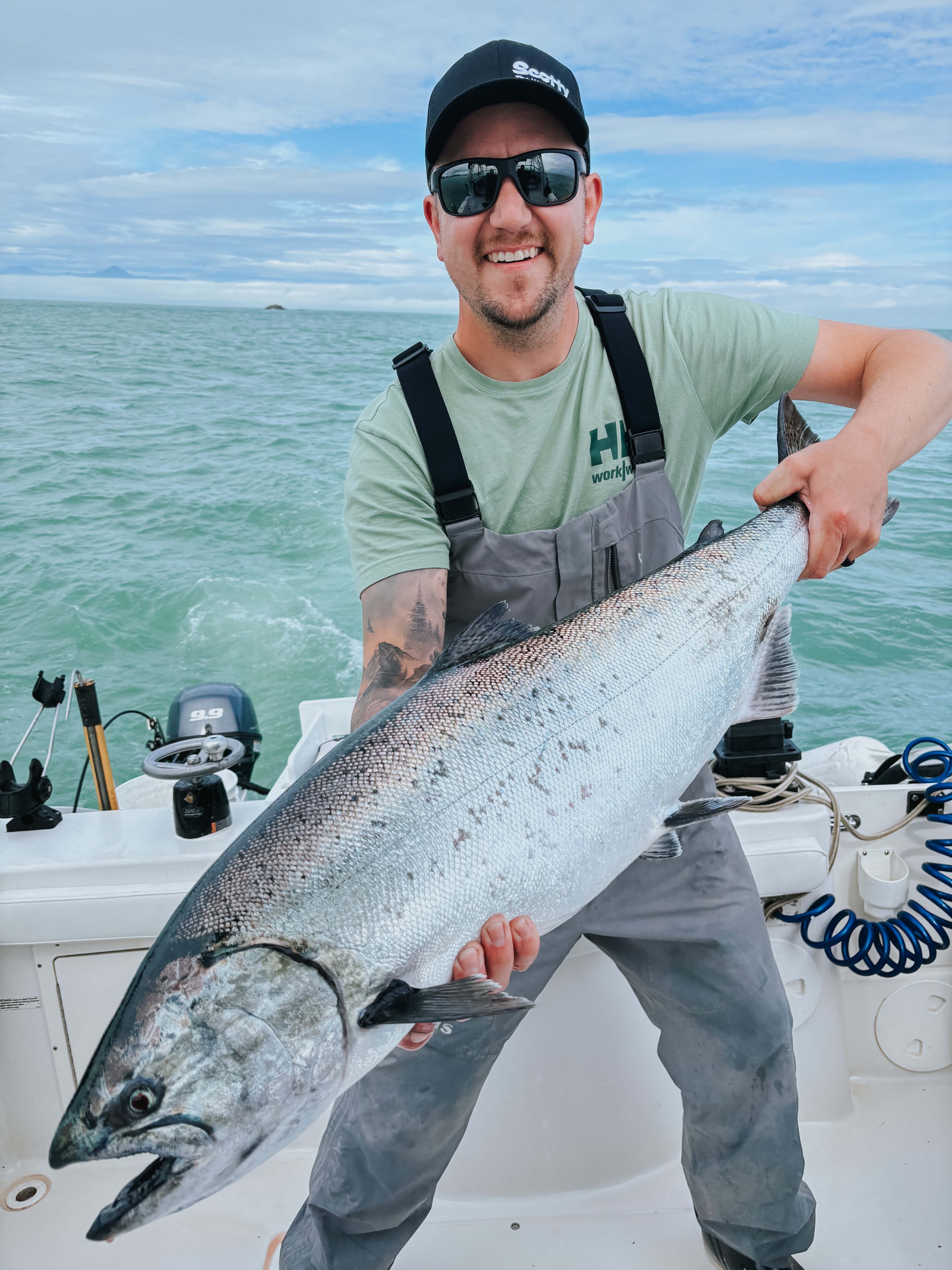 Smiling man wearing sunglasses and gray waders holding a large silver fish on a boat with ocean in the background.