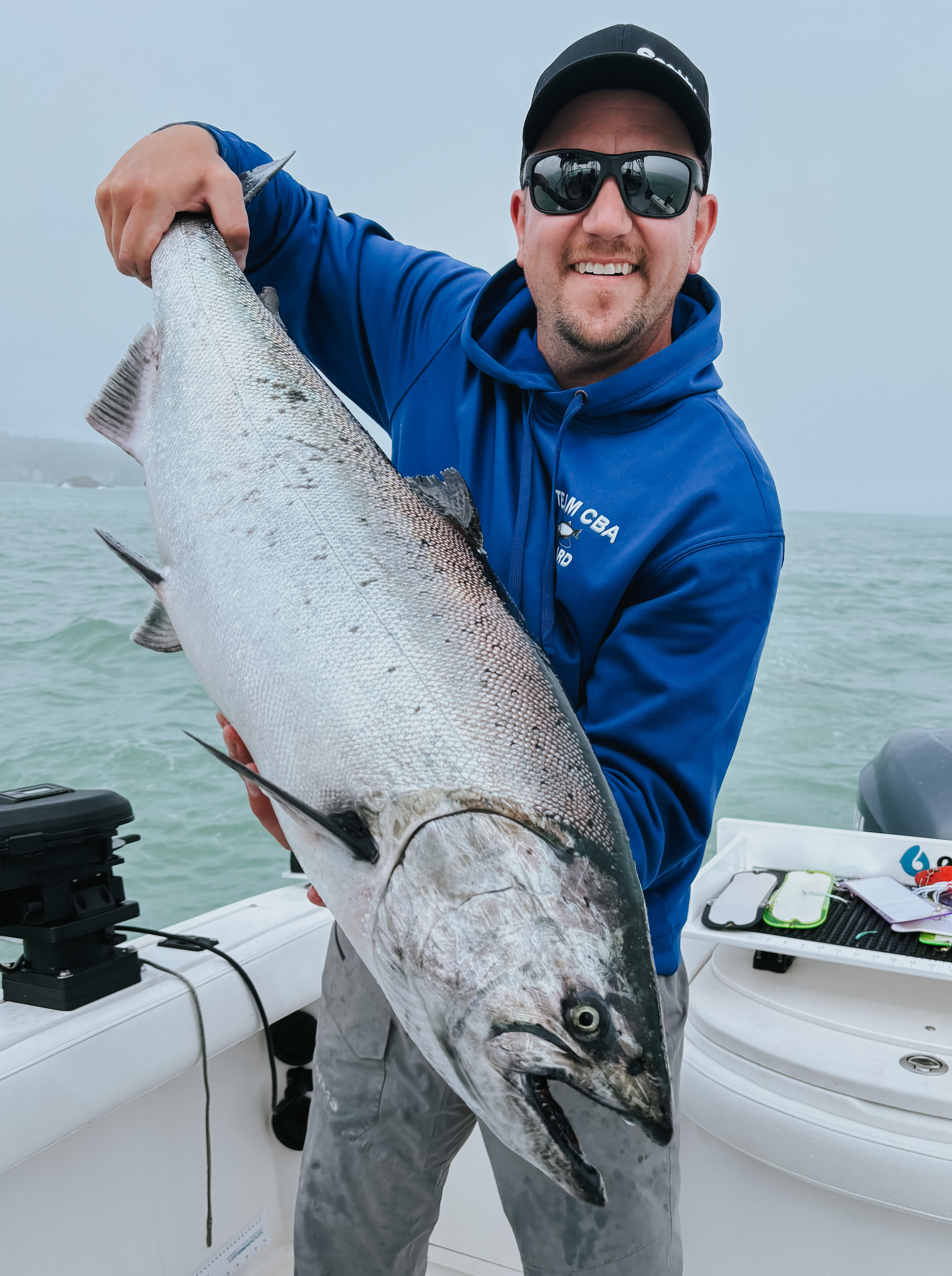 Smiling man wearing sunglasses and a blue hoodie holding a large salmon on a boat over water.