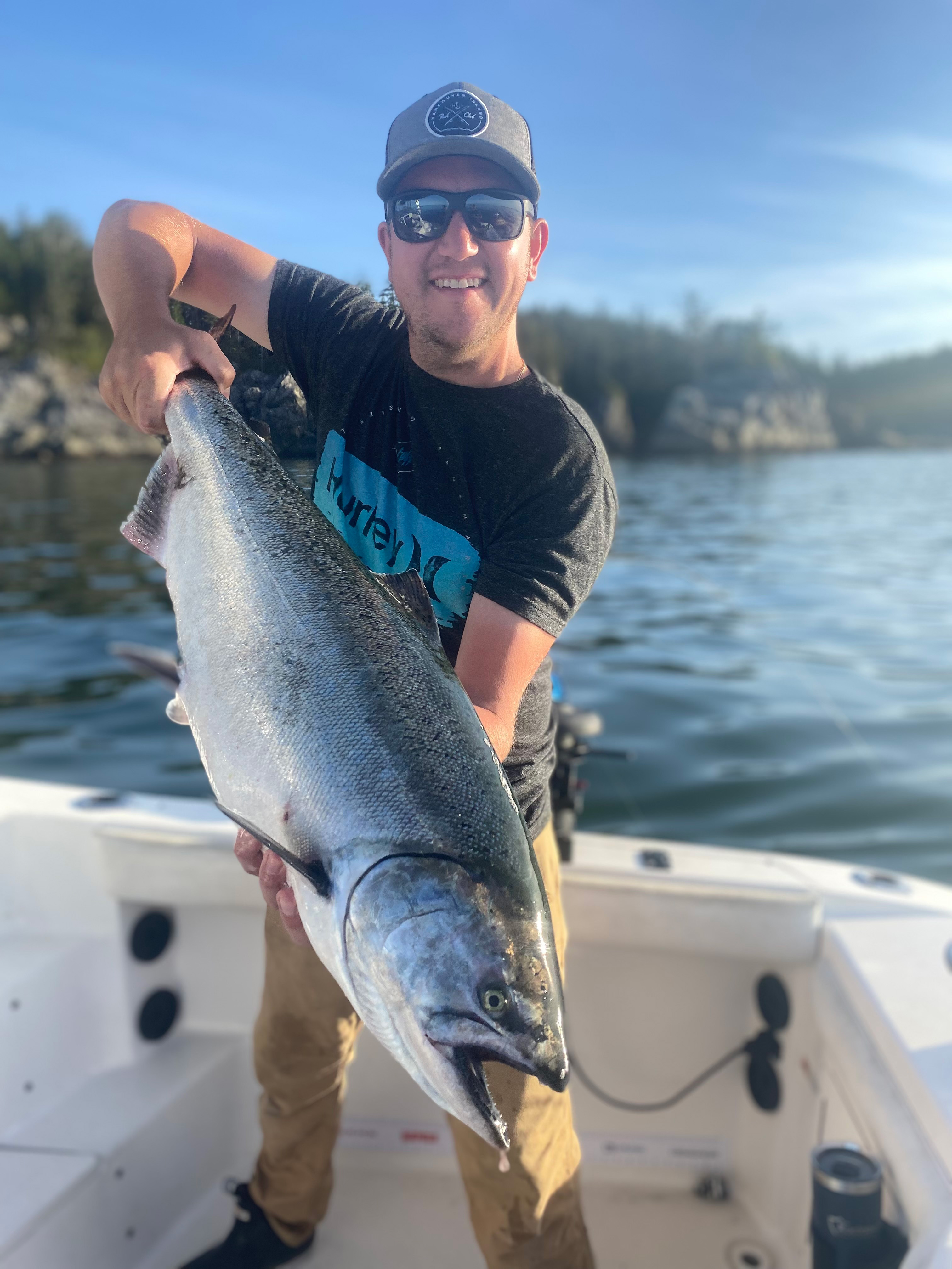 Smiling man wearing sunglasses and a cap holding a large salmon on a boat with water and rocky shore in the background.