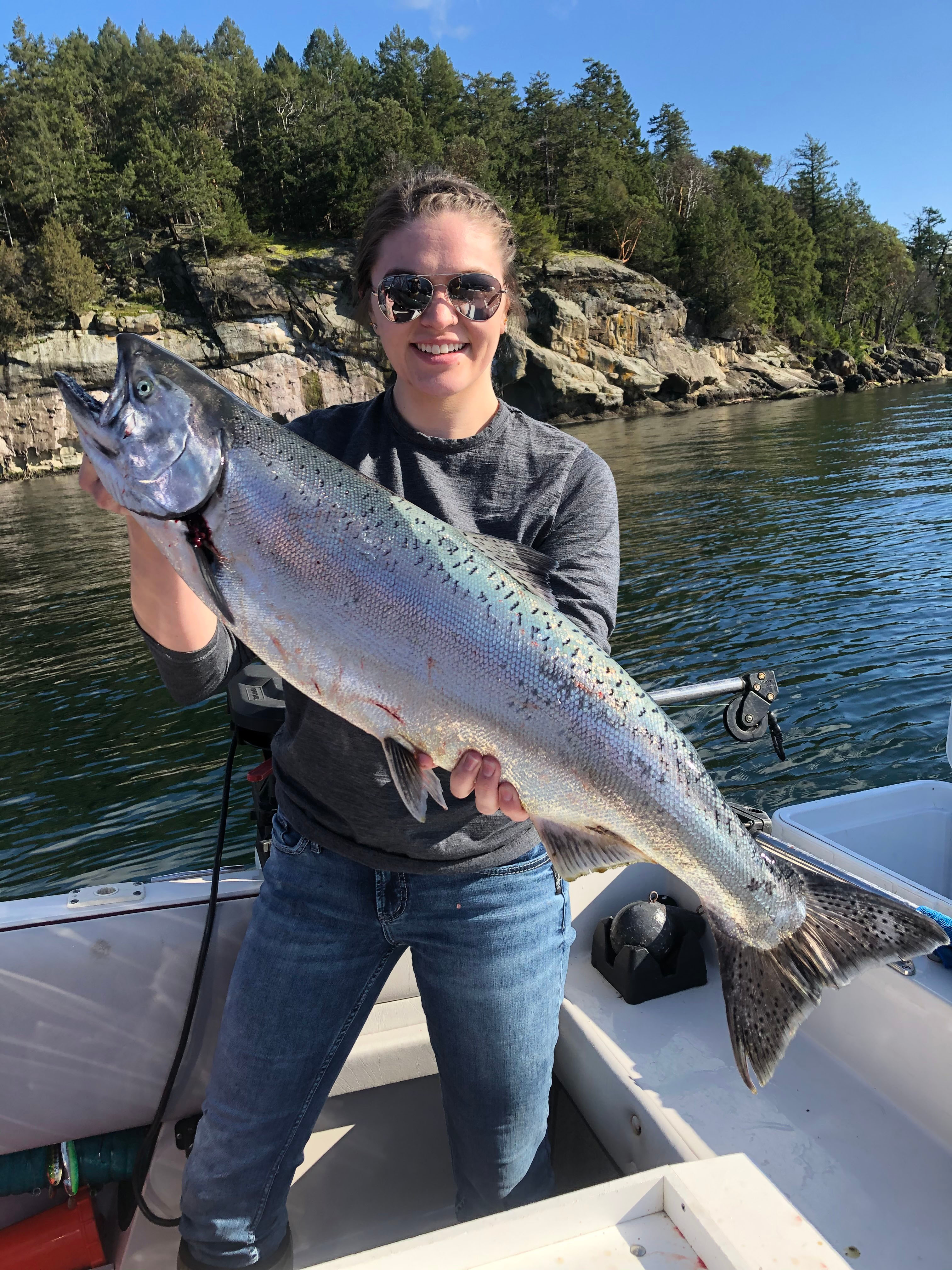 Woman wearing sunglasses holding a large salmon on a boat with forested rocky shoreline in the background.