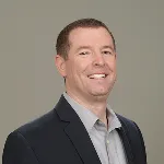 Smiling man wearing a dark blazer and light gray shirt against a neutral background.