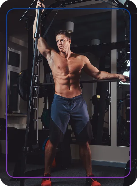 Shirtless muscular man in workout shorts holding gym equipment in a fitness center.