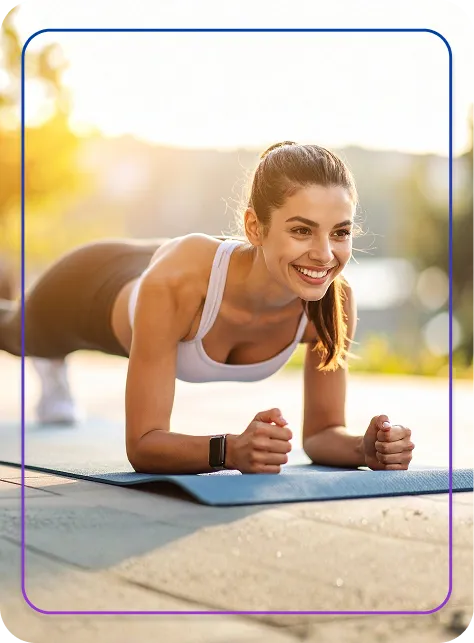 Smiling woman in workout attire holding a plank position on a blue exercise mat outdoors during sunrise or sunset.